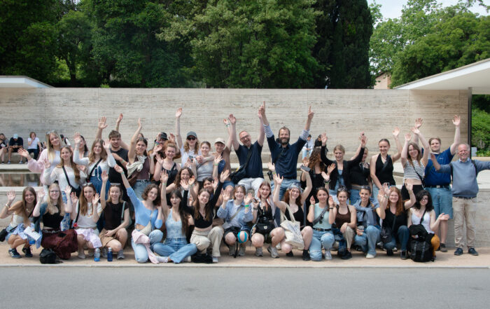 Eine große Gruppe von Menschen posiert gemeinsam im Freien, lächelnd und mit den Händen in der Luft winkend, mit Bäumen und einer Steinmauer, die schöne Architektur im Hintergrund zeigt. at Hochschule Coburg
