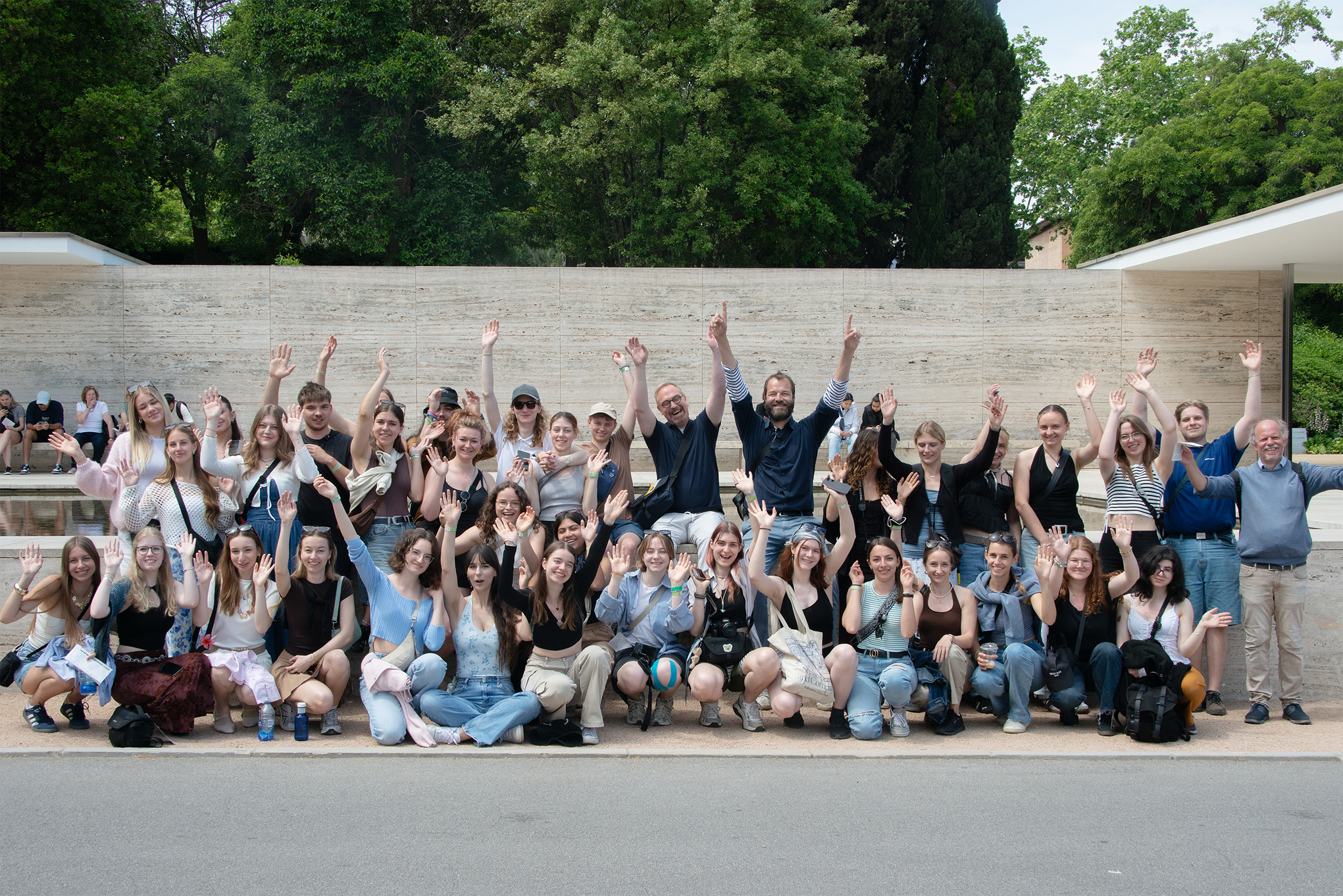Eine große Gruppe von Menschen posiert gemeinsam im Freien, lächelnd und mit den Händen in der Luft winkend, mit Bäumen und einer Steinmauer, die schöne Architektur im Hintergrund zeigt. at Hochschule Coburg