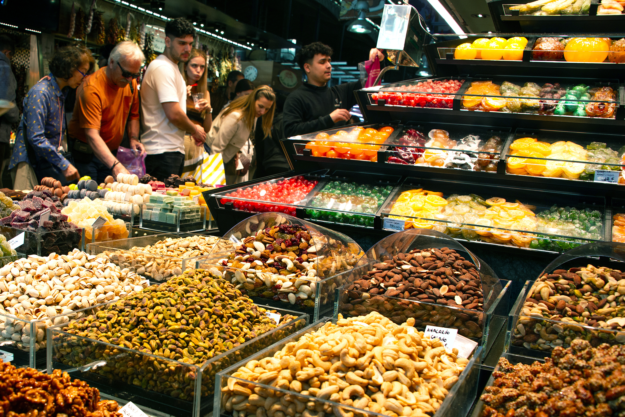 Käufer stöbern an einem belebten Marktstand in Barcelona, wo unter hellen Lichtern Tabletts mit Trockenfrüchten, Nüssen und bunten kandierten Bonbons ausgestellt sind. Die lebendige Szene spiegelt das einzigartige Design der Stadt wider und schafft eine einladende Atmosphäre. at Hochschule Coburg