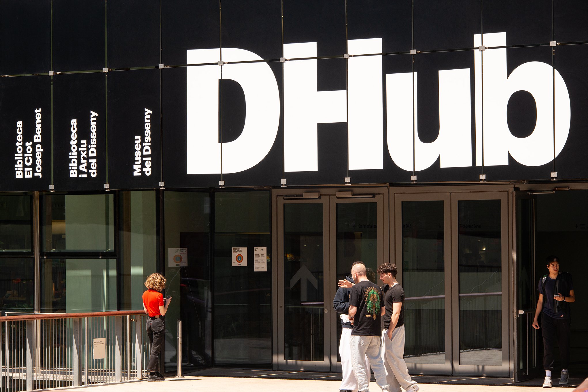 Menschen stehen und gehen in der Nähe des Eingangs eines modernen Gebäudes in Barcelona mit einem großen "DHUB"-Schild und einem kleineren vertikalen Text, auf dem Bibliotecca, Escola und Museu del Disseny aufgeführt sind, was eine beeindruckende Architektur und innovatives Design widerspiegelt. at Hochschule Coburg