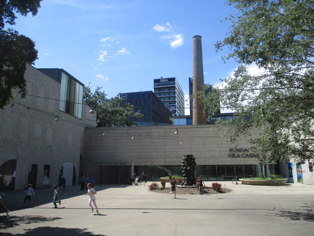 Kinder und Erwachsene spazieren in einem offenen Hof vor dem Gebäude der Fundació Vila Casas, in dem sich moderne Architektur mit markanten Designelementen vermischt und ein hoher Schornstein vor einem blauen Himmel mit vereinzelten Wolken emporragt. at Hochschule Coburg