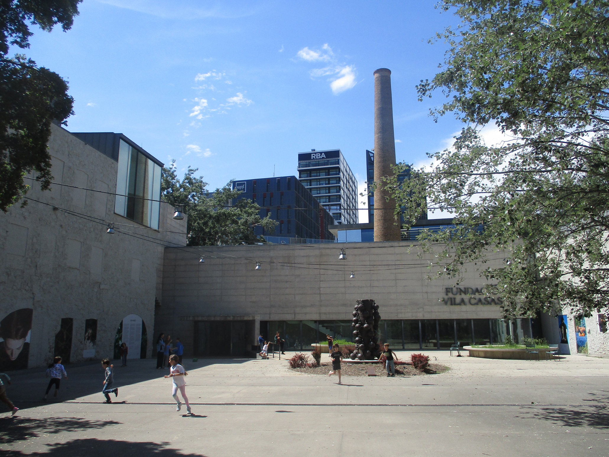 Kinder und Erwachsene spazieren in einem offenen Hof vor dem Gebäude der Fundació Vila Casas, in dem sich moderne Architektur mit markanten Designelementen vermischt und ein hoher Schornstein vor einem blauen Himmel mit vereinzelten Wolken emporragt. at Hochschule Coburg