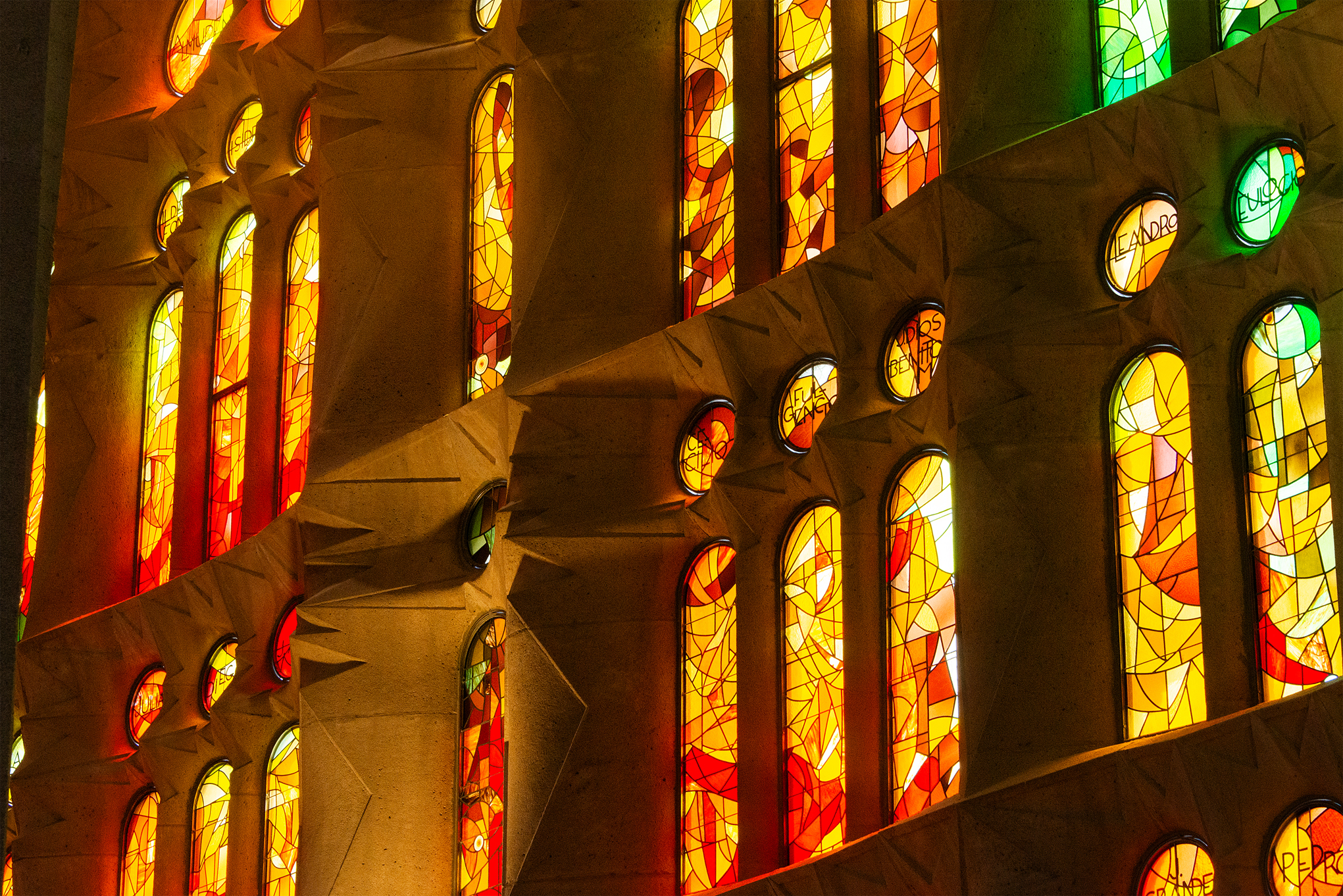 Bunte Glasfenster in einer strukturierten Steinwand, deren Design von der Architektur Barcelonas inspiriert ist, mit warmen Gelb-, Orange- und Rottönen in geometrischen Formen, die leuchtendes Licht in den Raum werfen. at Hochschule Coburg