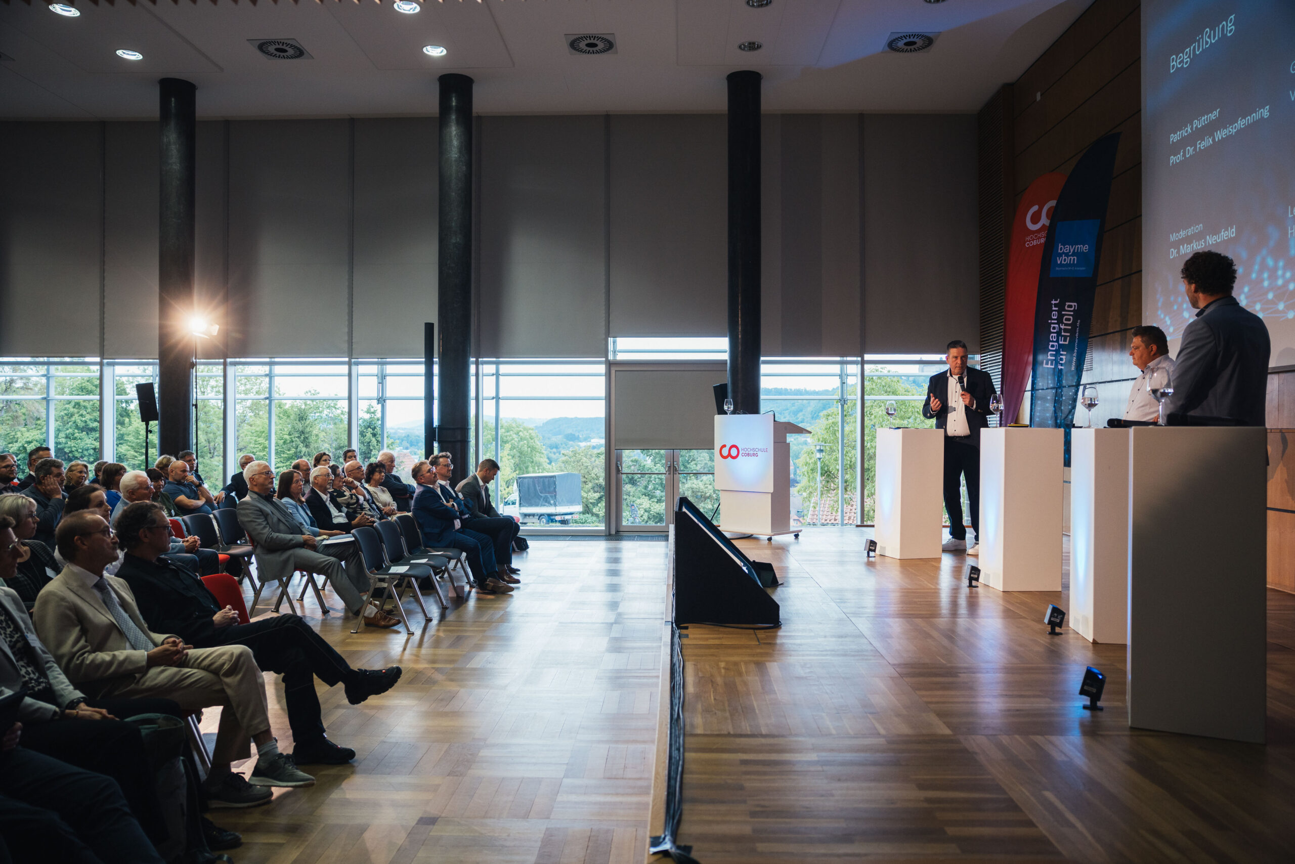 Eine Gruppe von Menschen sitzt in Reihen vor einer Bühne, auf der vier Redner an Podesten stehen und in einem modernen Konferenzraum mit großen Fenstern und Holzböden präsentieren und so den Austausch über Wissenschaft und Wirtschaft fördern. at Hochschule Coburg