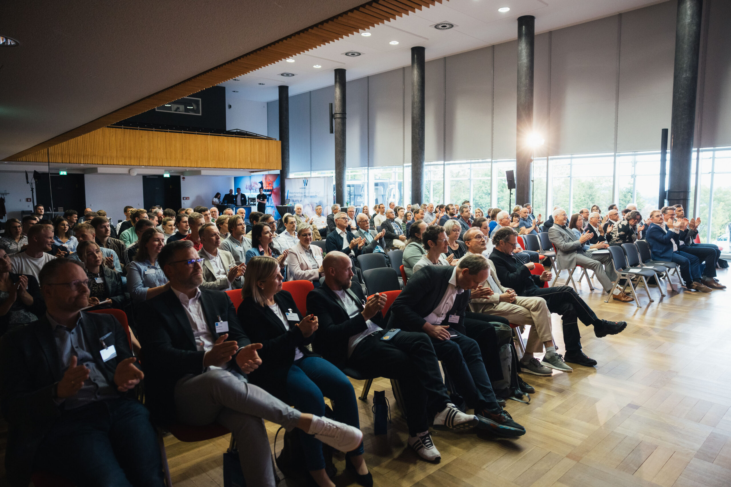 Eine große Gruppe von Menschen sitzt in Stuhlreihen in einem geräumigen, modernen Konferenzraum mit großen Fenstern, beobachtet aufmerksam eine Präsentation zu Wissenschaft und Wirtschaft und applaudiert ihr. Die Atmosphäre fördert Austausch und gemeinsames Lernen. at Hochschule Coburg