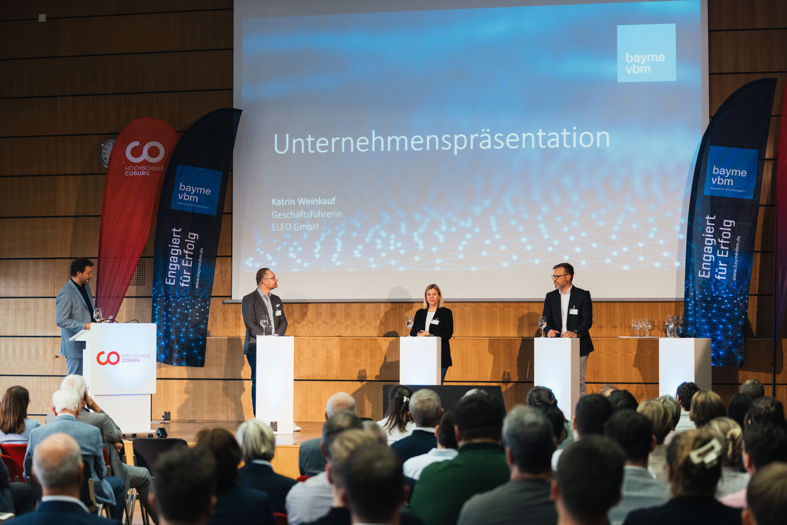 Eine Business-Präsentationsveranstaltung mit vier Rednern auf Podiumsplätzen, die sich an ein Publikum wenden. Auf der großen Leinwand sind „Unternehmenspräsentationen“ und Firmenlogos zu sehen, die den Austausch zwischen Wissenschaft und Wirtschaft hervorheben. Bunte Banner flankieren die Bühne. at Hochschule Coburg