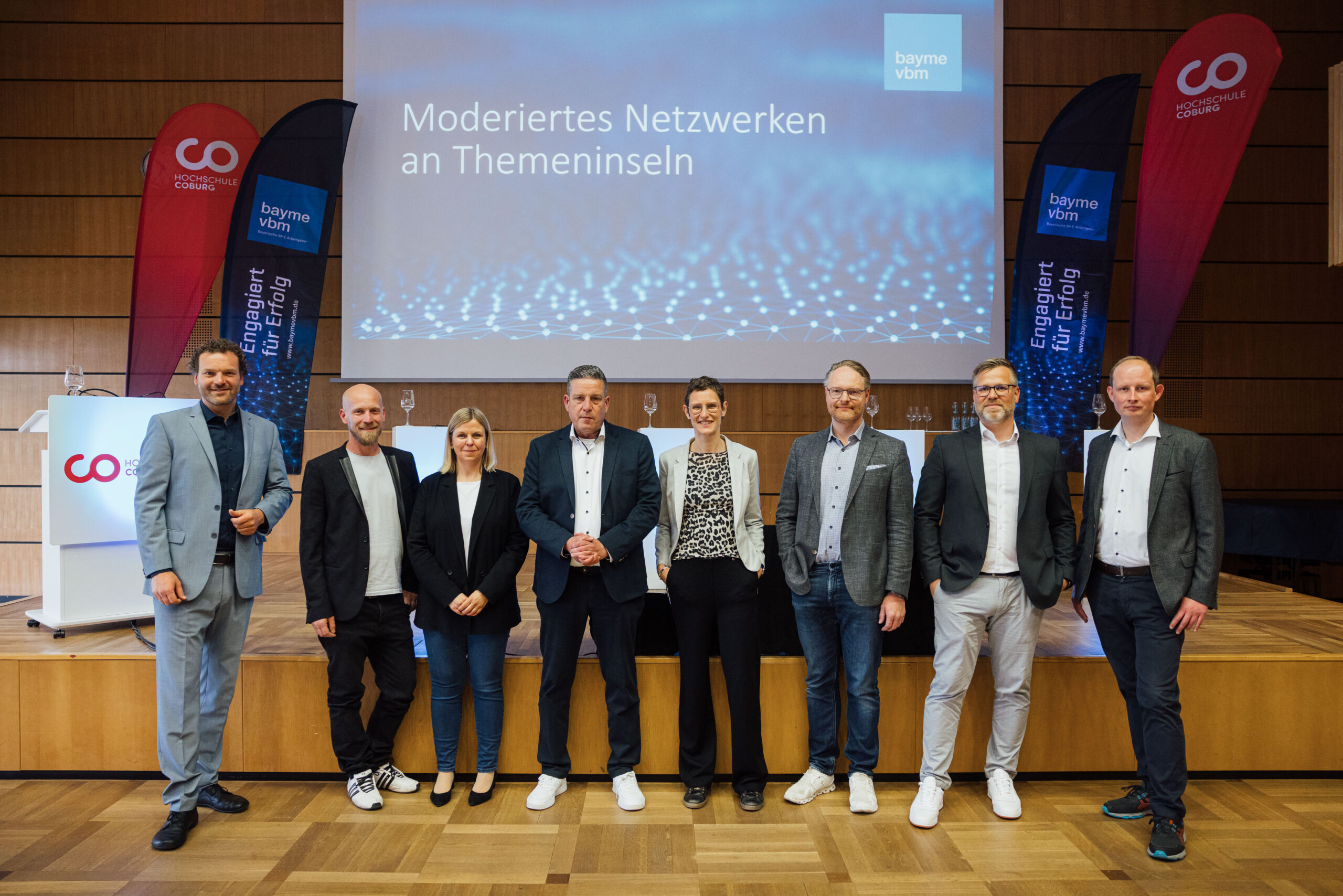 Neun Personen stehen in Business-Casual-Kleidung in einer Reihe auf einer Bühne, hinter ihnen zwei rote Banner und eine Leinwand mit der Aufschrift „Moderierte Netzwerke an Themeninseln“, die spannende Einblicke in Wirtschaft und Wissenschaft bieten. at Hochschule Coburg