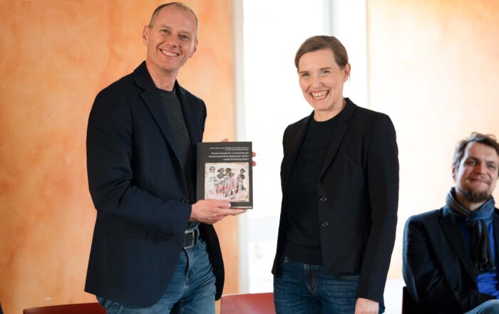 Zwei Personen stehen im Innenbereich und lächeln in die Kamera. Die linke Person hält ein Buch, die andere steht daneben. Beide tragen dunkle Blazer und Jeans – was ihre Verbundenheit zur Sozialen Arbeit widerspiegelt. Eine weitere Person sitzt im Hintergrund, etwas unscharf. at Hochschule Coburg