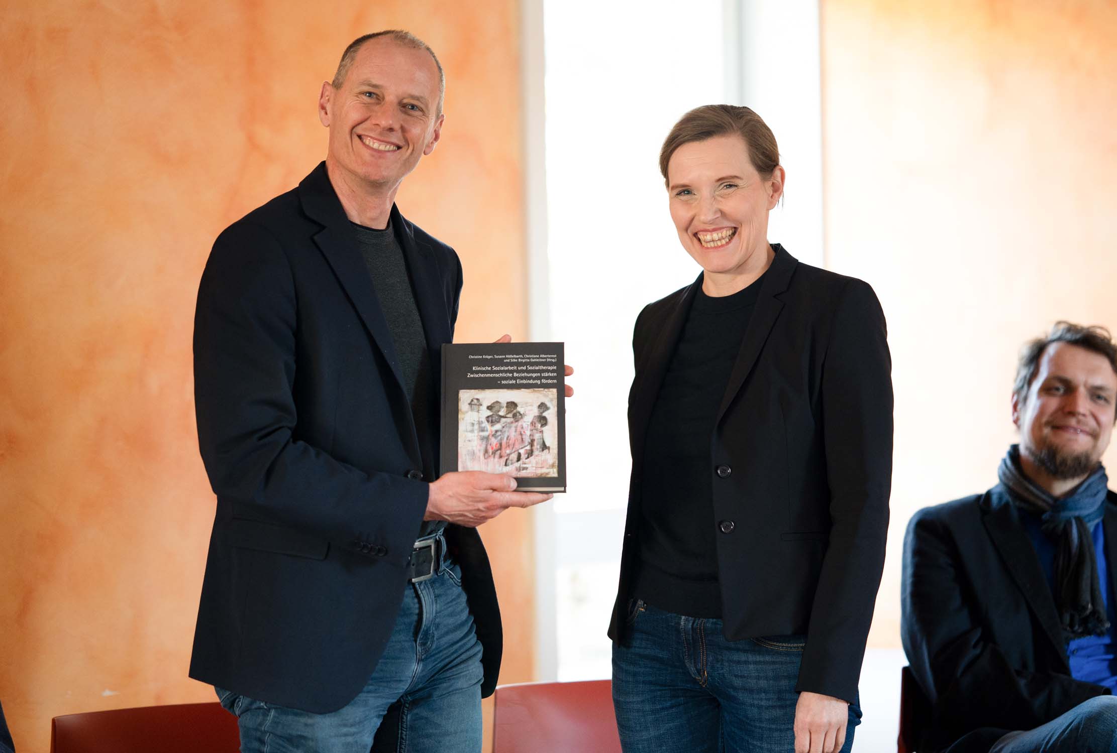 Zwei Personen stehen im Innenbereich und lächeln in die Kamera. Die linke Person hält ein Buch, die andere steht daneben. Beide tragen dunkle Blazer und Jeans – was ihre Verbundenheit zur Sozialen Arbeit widerspiegelt. Eine weitere Person sitzt im Hintergrund, etwas unscharf. at Hochschule Coburg