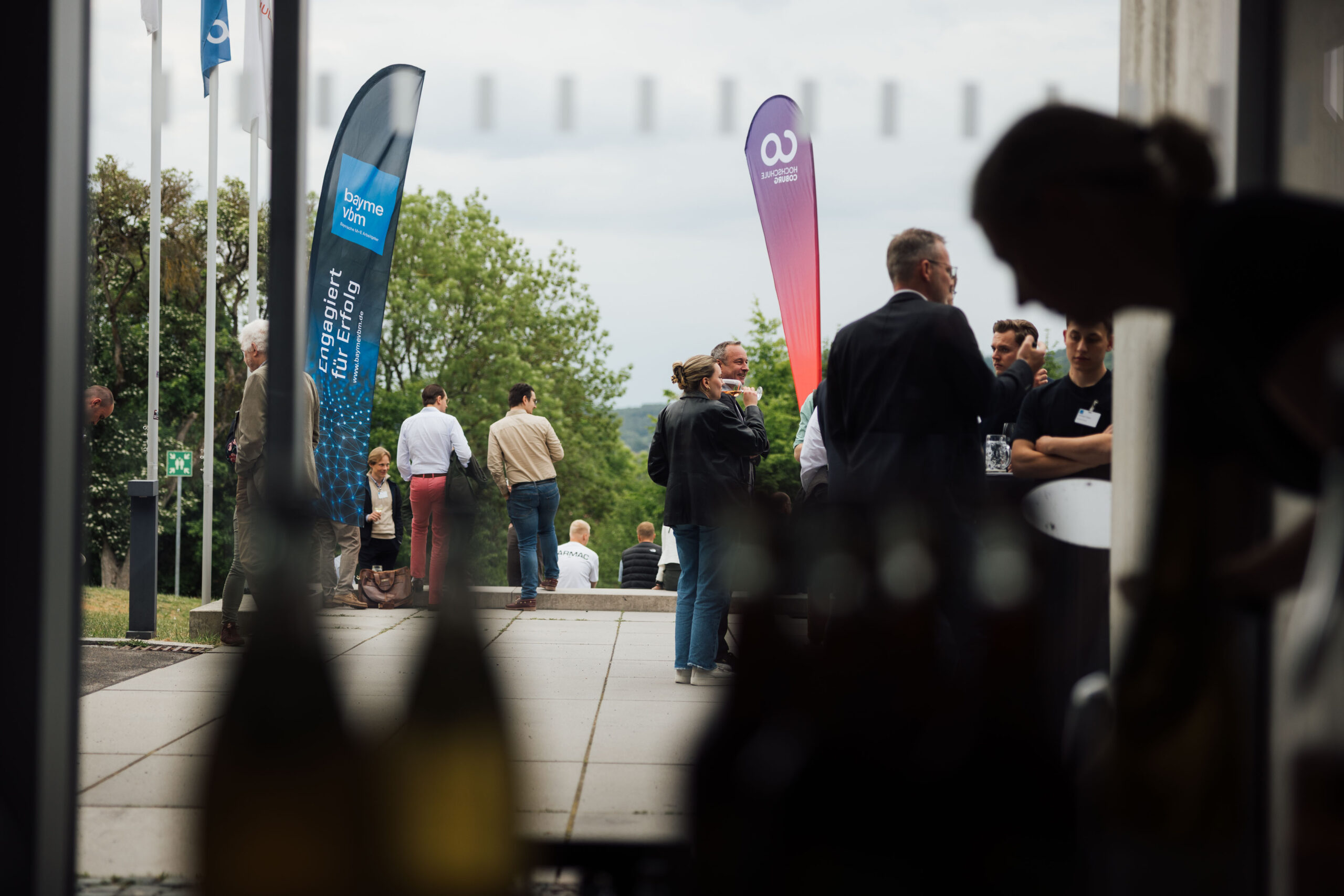 Menschen treffen sich im Freien bei einer Veranstaltung mit bunten Bannern, tauschen sich aus und diskutieren angeregt. Im Vordergrund sind Flaschen zu sehen, Bäume und ein bewölkter Himmel bilden den Hintergrund. at Hochschule Coburg