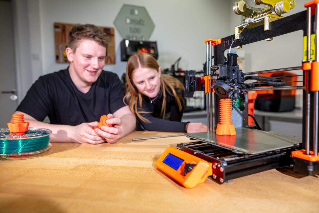 Zwei Menschen lächeln, während sie einem 3D-Drucker beim Erstellen eines orangefarbenen Objekts zusehen. In diesem hellen, modernen Arbeitsbereich zeigen Filamentspulen und fertige Drucke den Innovationsgeist der Wirtschaftsinformatik 2.0 in Aktion. at Hochschule Coburg