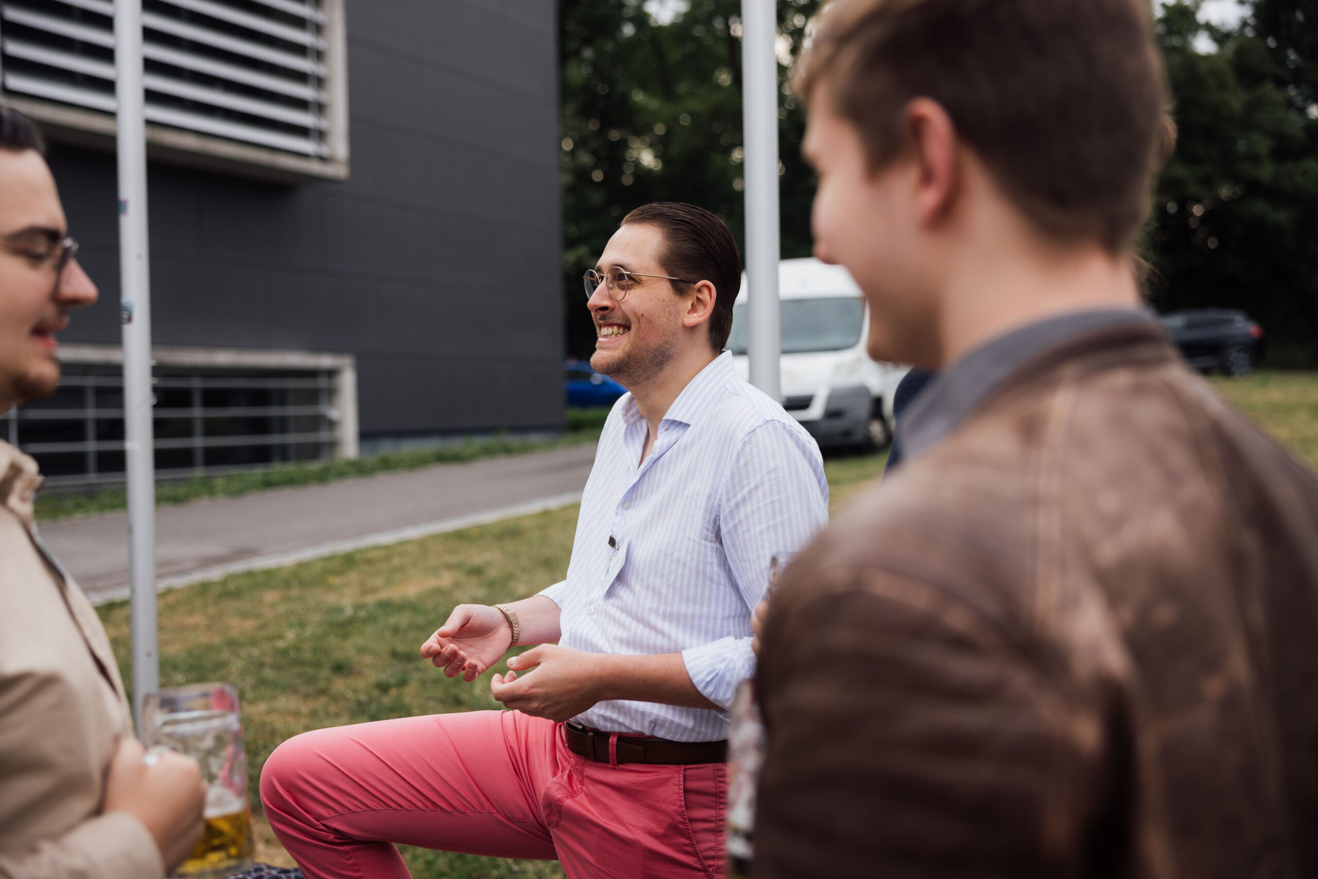 Drei Männer stehen im Freien, unterhalten sich und lächeln. Ein Mann in der Mitte trägt ein weißes Hemd und eine rosa Hose. Die Szene bietet Einblicke in die Wirtschaft vor einer Kulisse aus Gras, einem Gebäude und einem geparkten Lieferwagen. at Hochschule Coburg
