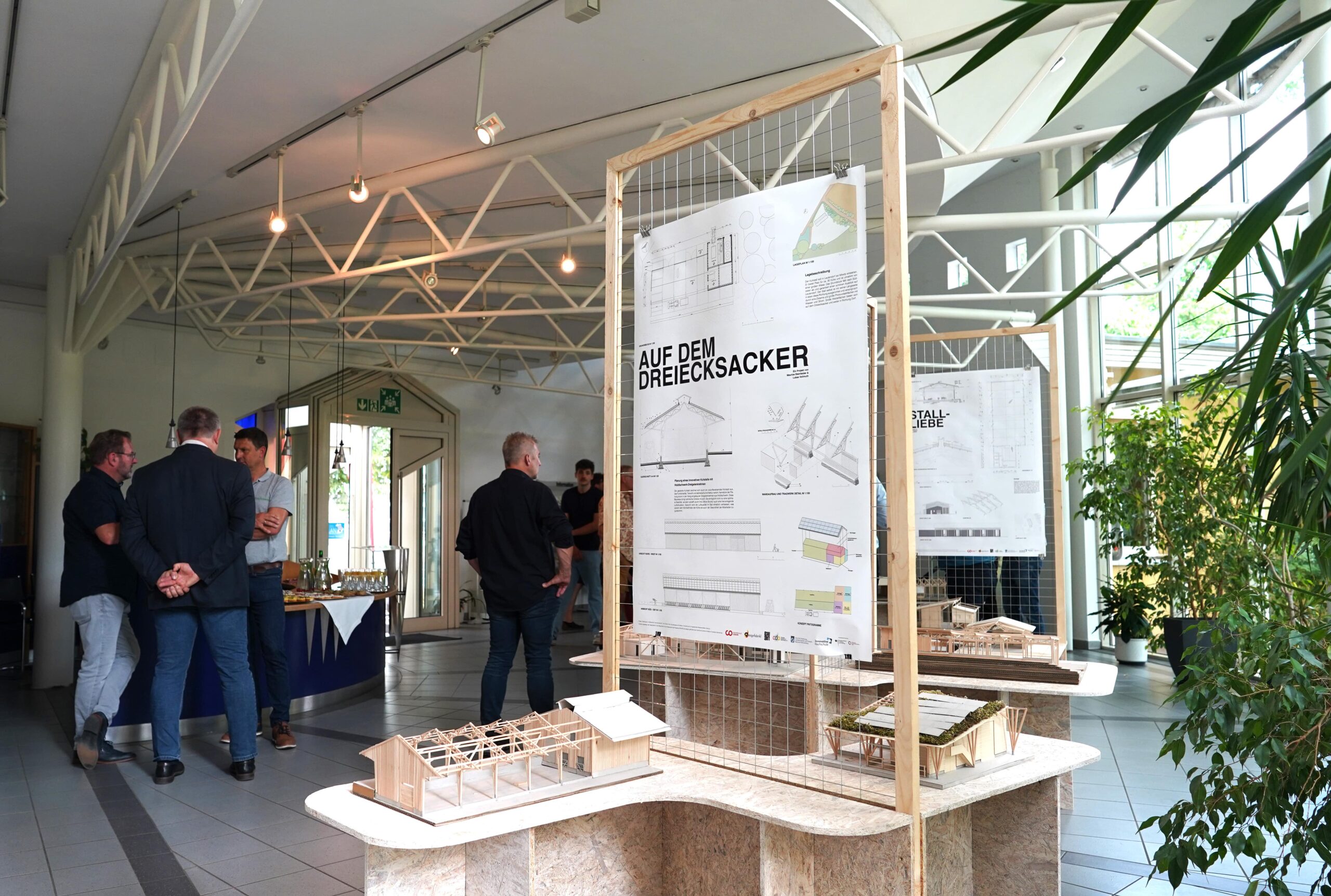 Ausstellungsraum mit ausgestellten Architekturmodellen und Projektplakaten; im Hintergrund sind mehrere Personen versammelt und unterhalten sich unter einer weißen Balkendecke, wobei Pflanzen und natürliches Licht sichtbar sind. at Hochschule Coburg