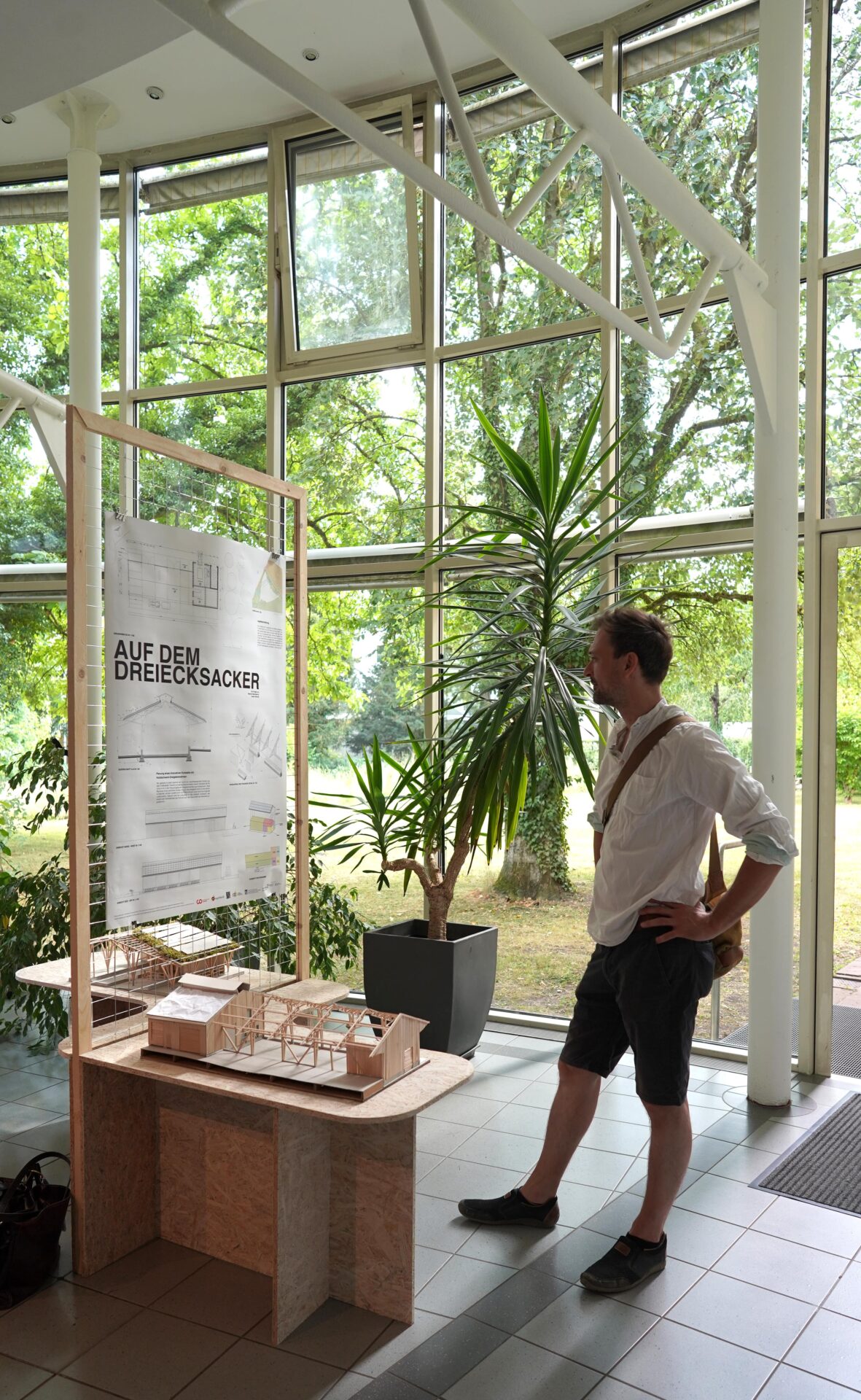 Ein Mann im weißen Hemd steht mit den Händen in den Hüften und betrachtet eine Architekturausstellung mit Modellen und einem Plakat mit der Aufschrift "AUF DEM DREIECKSACKER" in einem hellen, modernen Raum mit großen Fenstern und Zimmerpflanzen. at Hochschule Coburg