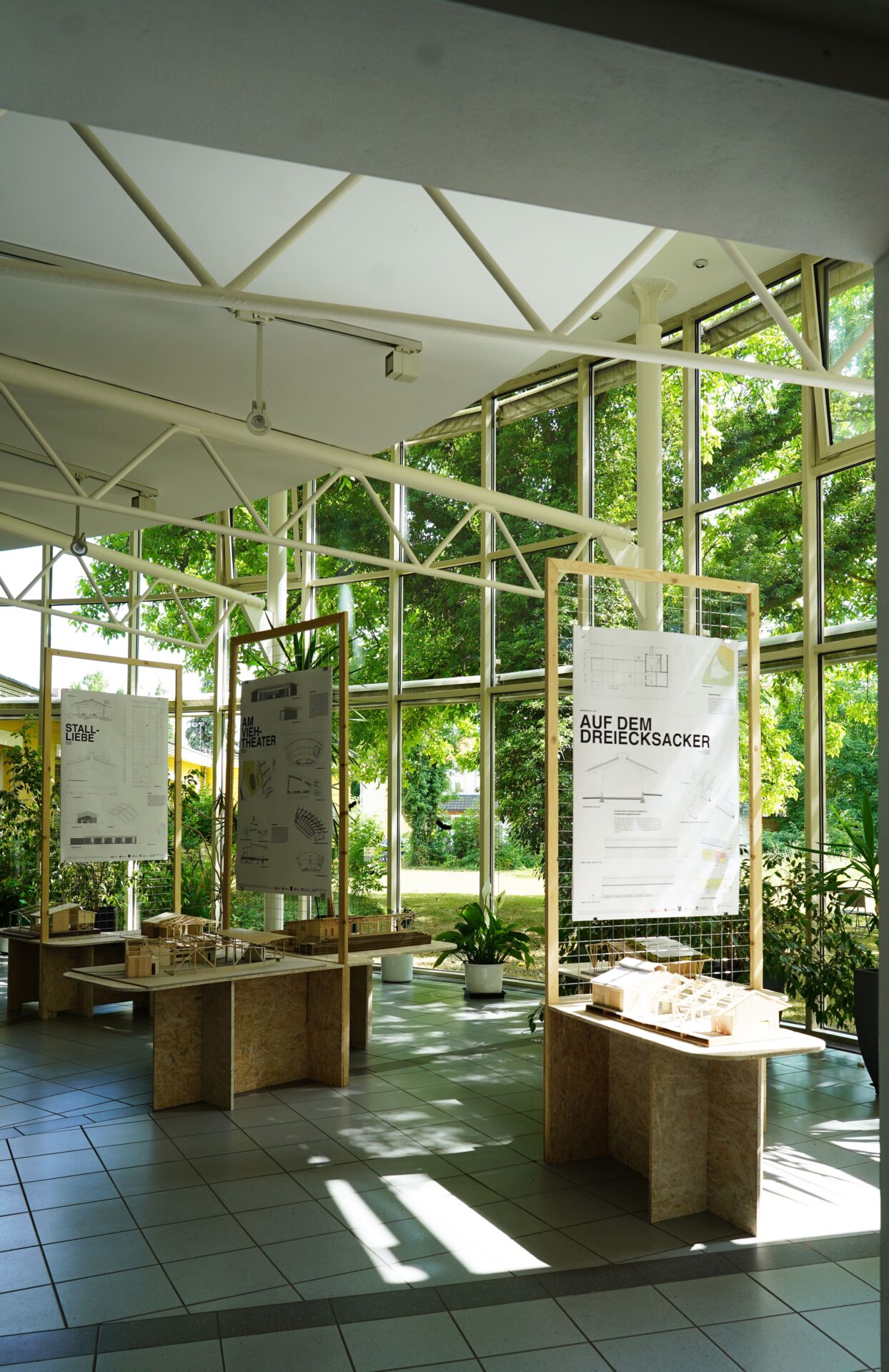 Ein sonnendurchfluteter Ausstellungsraum mit großen Fenstern, in dem Architekturplakate und kleine Gebäudemodelle auf Tischen ausgestellt sind, umgeben von Grünpflanzen und Bäumen, die von außen sichtbar sind. at Hochschule Coburg