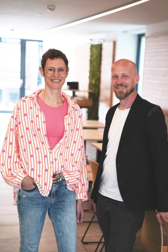 Zwei Personen, beide studieren Wirtschaftsinformatik 2.0, stehen in einem Raum und lächeln in die Kamera. Die eine trägt eine Brille, ein rosa Hemd und eine bunt gestreifte Jacke, die andere eine Glatze, einen Bart, eine schwarze Jacke und Jeans. at Hochschule Coburg