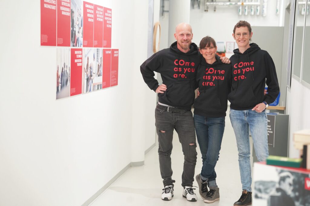 Drei Menschen stehen in einem hell erleuchteten Flur und lächeln in die Kamera. Sie tragen passende schwarze Kapuzenpullover mit der Aufschrift „Come as you are“, an der Wand neben ihnen hängen rote Plakate, die für Wirtschaftsinformatik 2.0 werben. at Hochschule Coburg