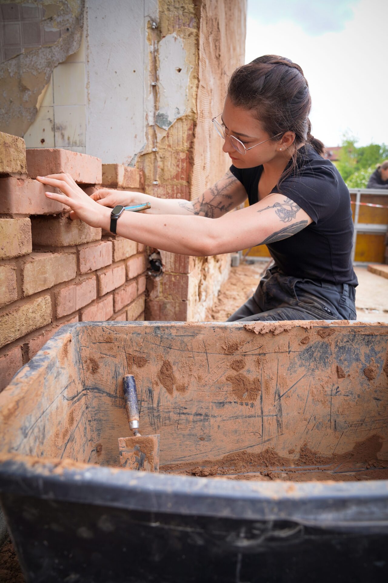 Eine Frau mit Brille und schwarzem Hemd legt vorsichtig Ziegelsteine beim Mauerbau im Freien. Sie arbeitet mit Mörtelwanne und Kelle. Die Mauer hinter ihr weist einige freiliegende, verwitterte Oberflächen auf. at Hochschule Coburg