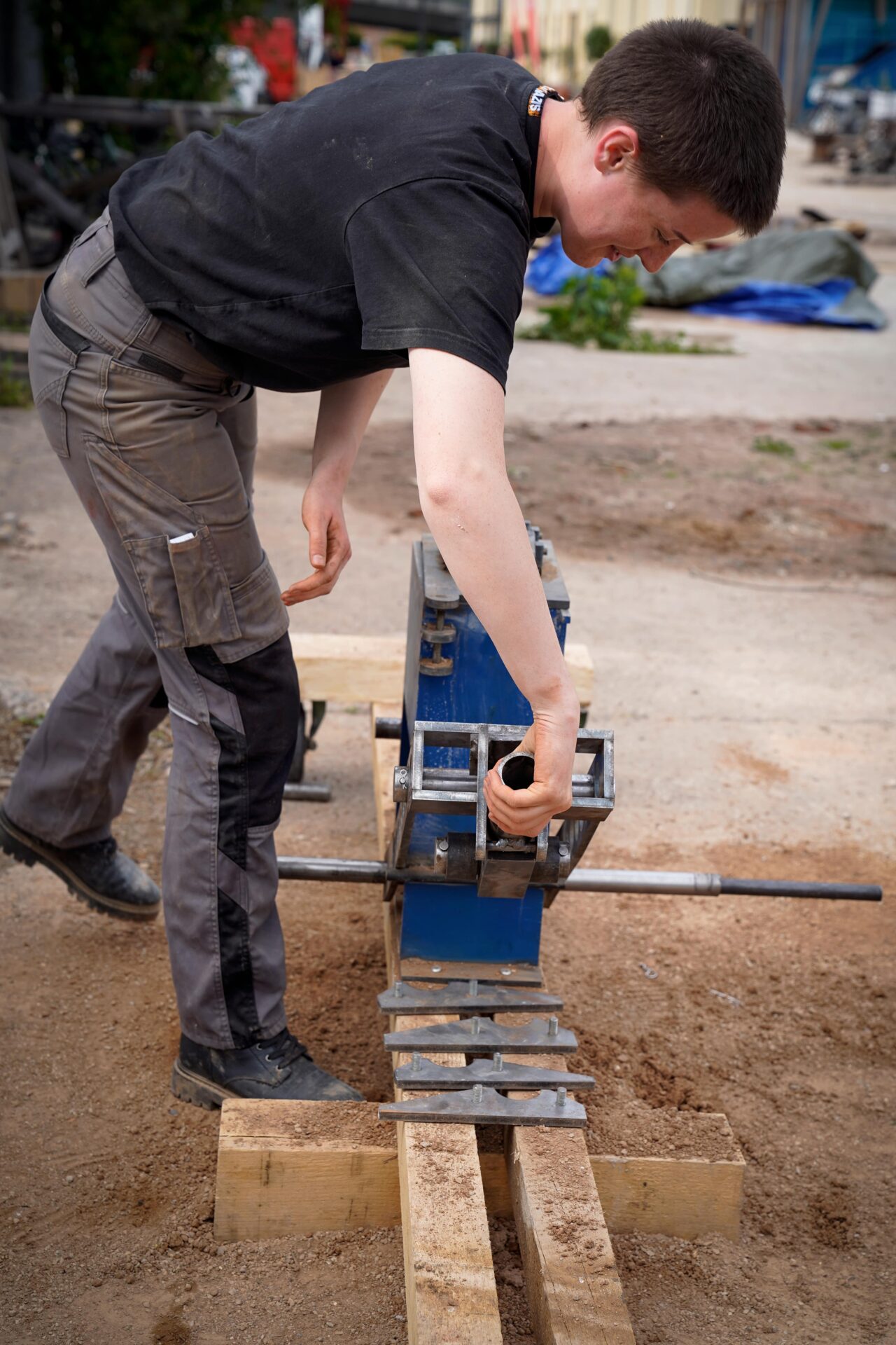 Eine Person in Arbeitskleidung beugt sich im Freien über ein blaues Metallwerkzeug und schneidet oder formt damit eine auf Holzstützen liegende Metallstange. Die Szene scheint auf einer Baustelle oder in einer Werkstatt zu spielen. at Hochschule Coburg
