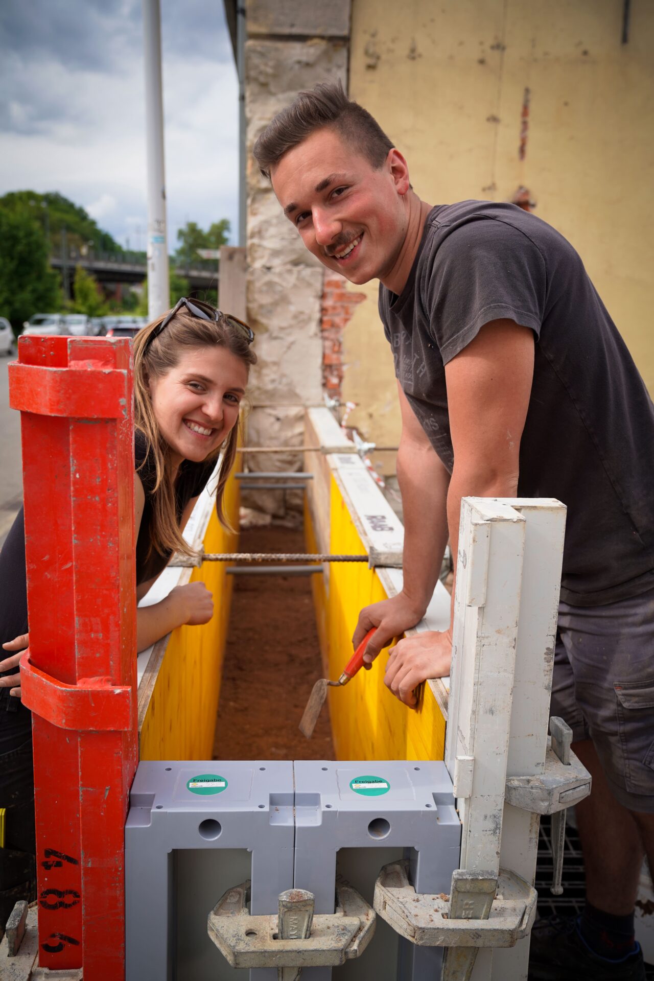Ein lächelnder Mann und eine lächelnde Frau arbeiten gemeinsam auf einer Baustelle. Sie beugen sich über einen mit gelb-weißen Absperrungen gesäumten Graben. Der Mann hält ein Werkzeug in der Hand, und beide scheinen mit ihrer Arbeit beschäftigt zu sein. at Hochschule Coburg