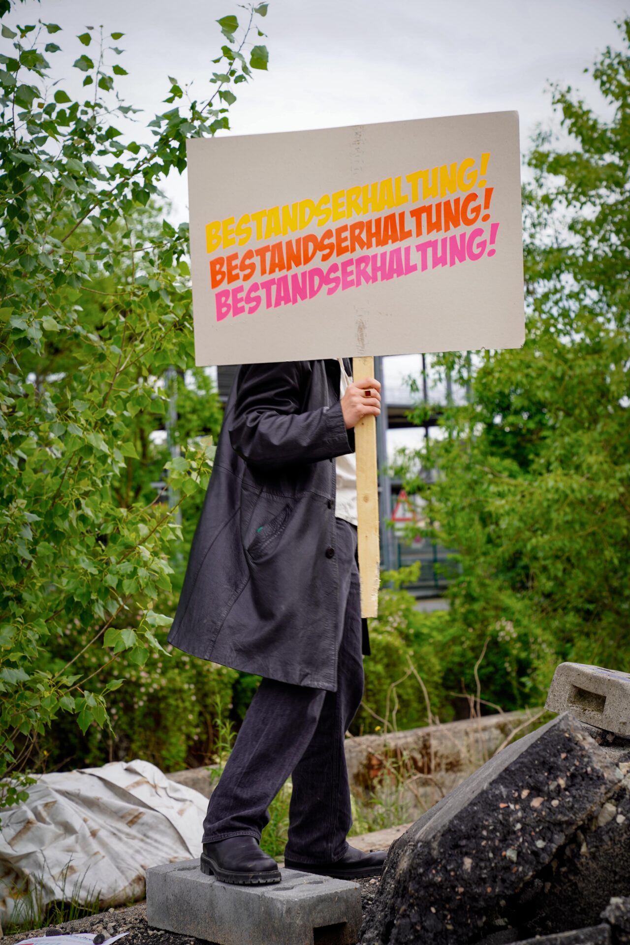 Eine Person steht im Freien auf Betonblöcken und hält ein Schild mit der Aufschrift „BESTANDSERHALTUNG!“ in leuchtend gelben und rosa Buchstaben. Ihr Gesicht ist durch das Schild verdeckt, im Hintergrund ist Grün zu sehen. at Hochschule Coburg
