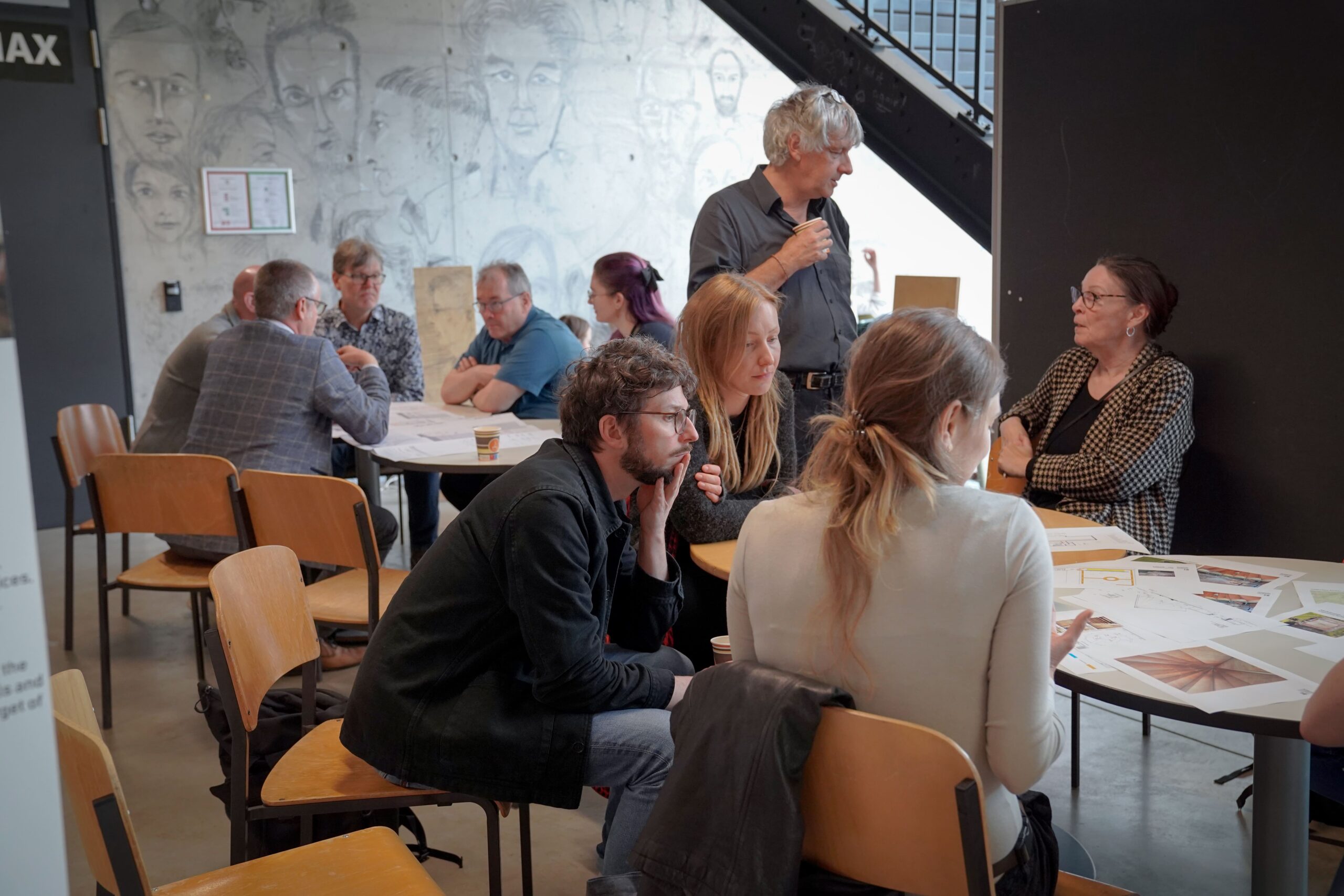 Mehrere Personen sitzen in einem hellen, modernen Raum an Tischen, diskutieren und lesen Dokumente. Eine Person steht und spricht mit einer sitzenden Frau. Im Hintergrund sind Bleistiftskizzen an der Wand zu sehen. at Hochschule Coburg