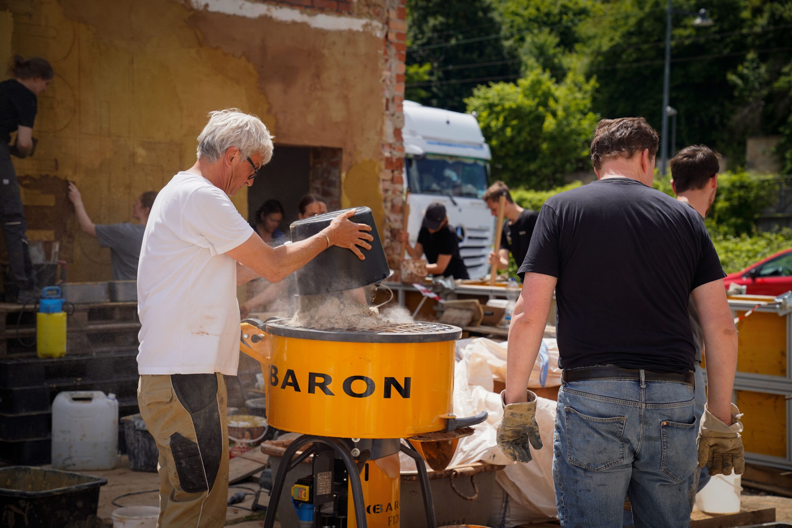 Mehrere Personen arbeiten im Freien und mischen Materialien in einer großen gelben Baron-Maschine. Eine Person schüttet Pulver aus einem Eimer in den Mischer, während andere in der Nähe Materialien vorbereiten und handhaben. Im Hintergrund sind ein LKW und Grünflächen zu sehen. at Hochschule Coburg