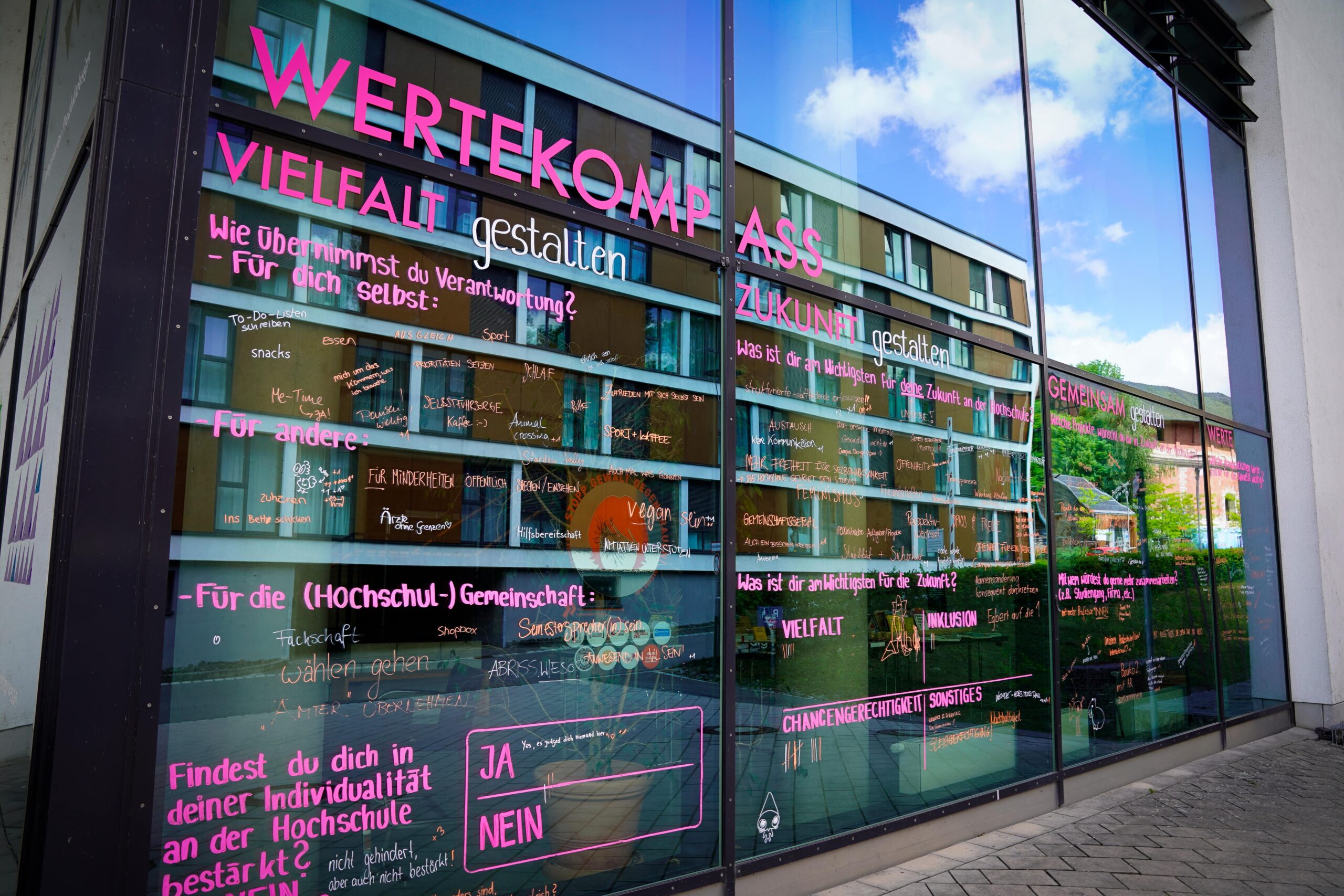 Auf den Glasfenstern sind bunte deutsche Texte zu Werten, Vielfalt und Gemeinschaft zu sehen, die mit verschiedenfarbigen Filzstiften geschrieben sind. In den Glasscheiben spiegeln sich Gebäude und Himmel. at Hochschule Coburg