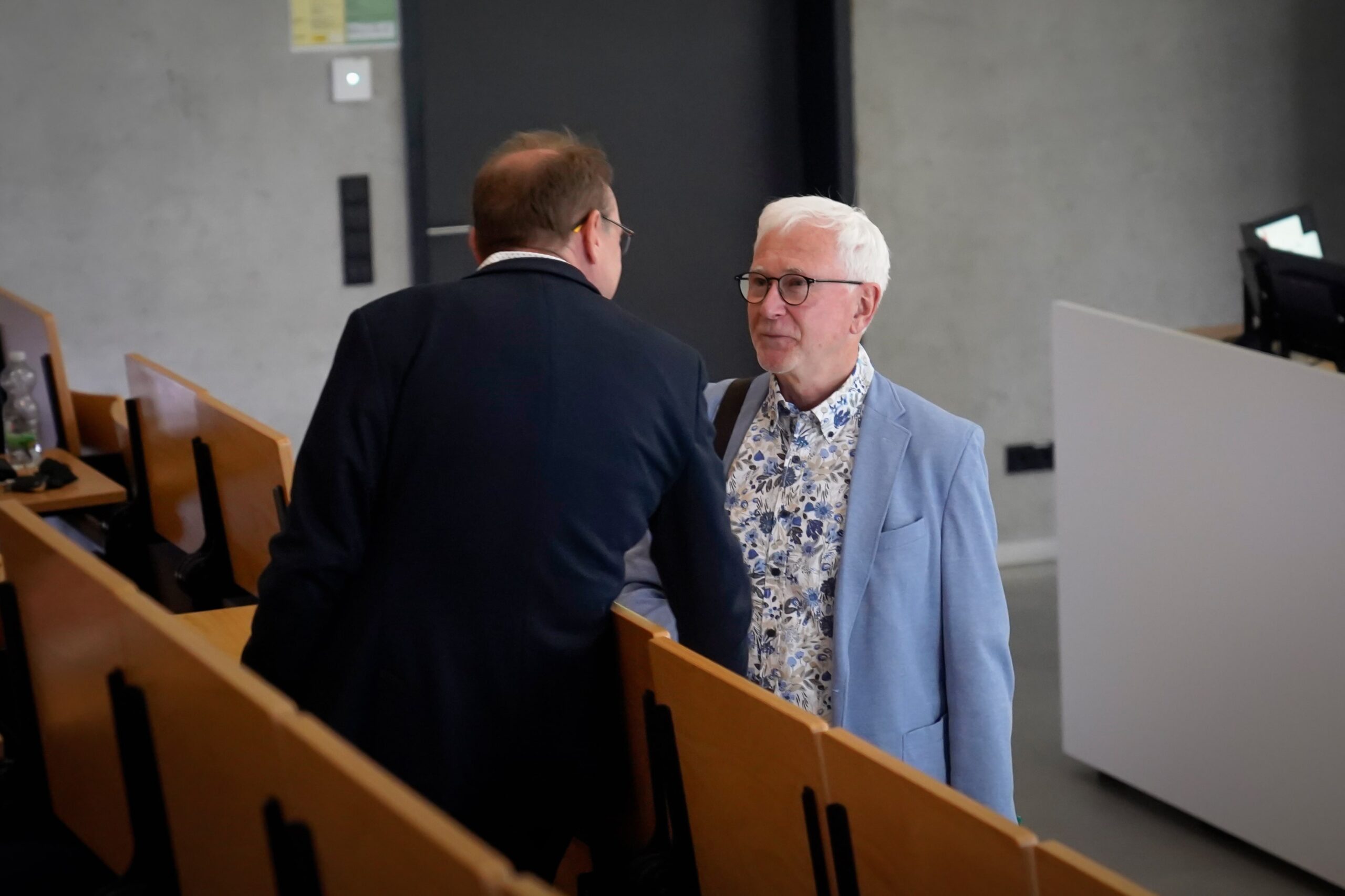 Zwei Männer unterhalten sich in einem Hörsaal. Ein Mann steht und trägt eine hellblaue Jacke und ein geblümtes Hemd, der andere sitzt und blickt abgewandt, beide sind von Holztischen umgeben. at Hochschule Coburg