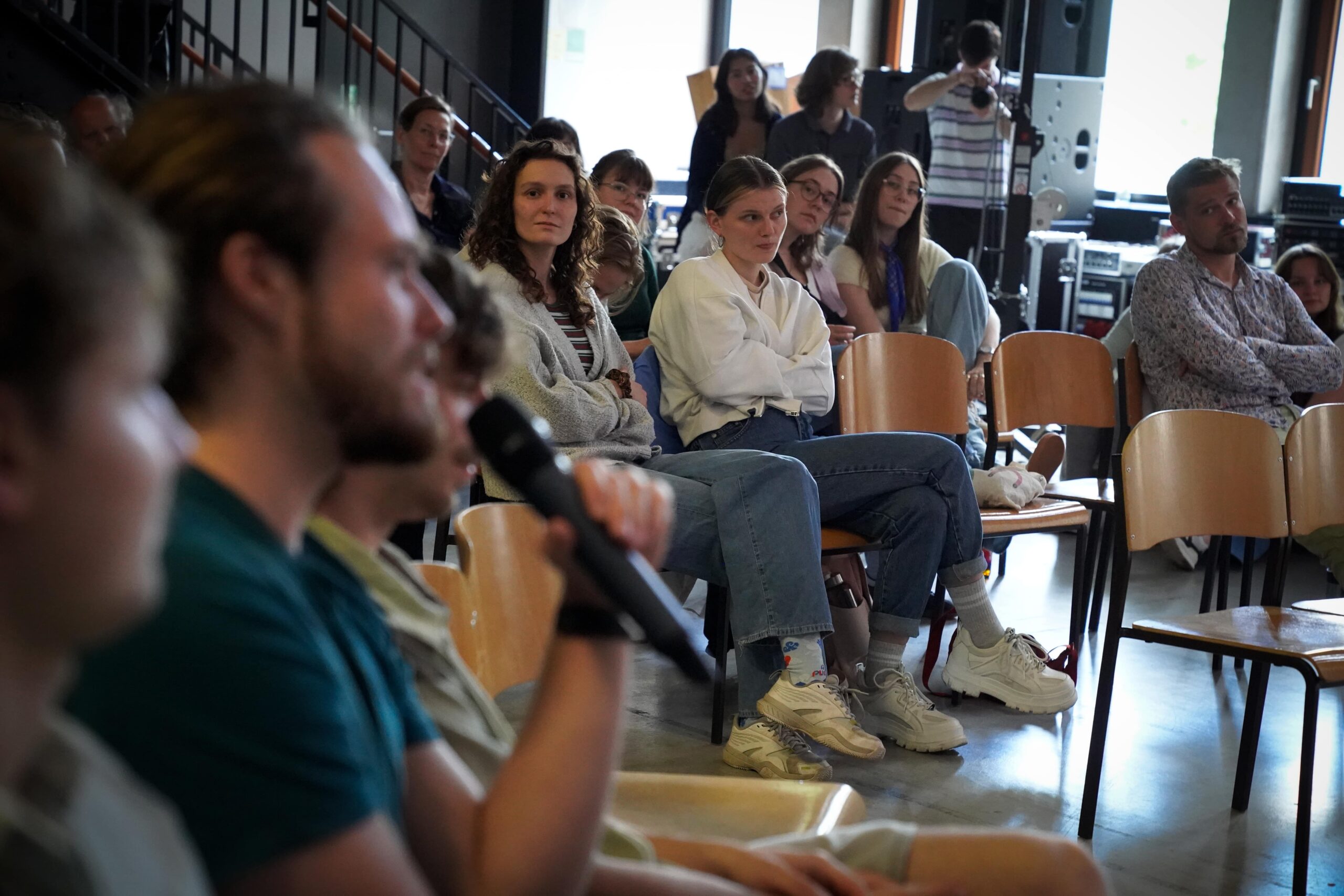 Eine Gruppe von Menschen sitzt auf Stühlen in einem Raum. Einige hören aufmerksam zu, während eine Person im Vordergrund in ein Mikrofon spricht. Es scheint sich um eine informelle Veranstaltung oder Diskussion zu handeln. at Hochschule Coburg
