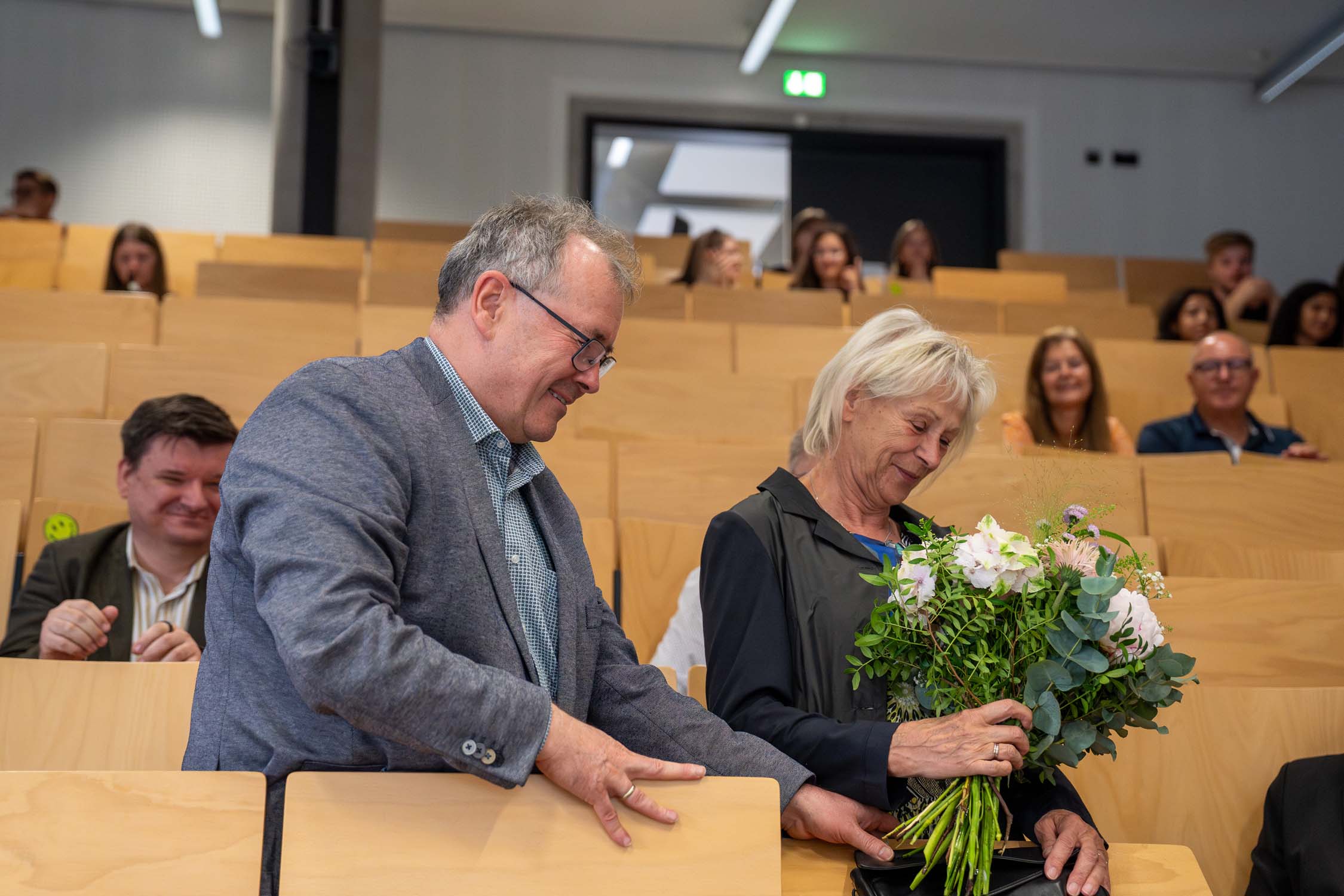 Ein Mann hilft einer älteren Frau mit einem Blumenstrauß in einem Hörsaal beim Aufstehen, vermutlich während der Verleihung der Kurbasik- und Hanses-Preise im Fach Bauingenieurwesen. Beide lächeln herzlich und teilen einen feierlichen Moment inmitten der sitzenden Gäste. at Hochschule Coburg