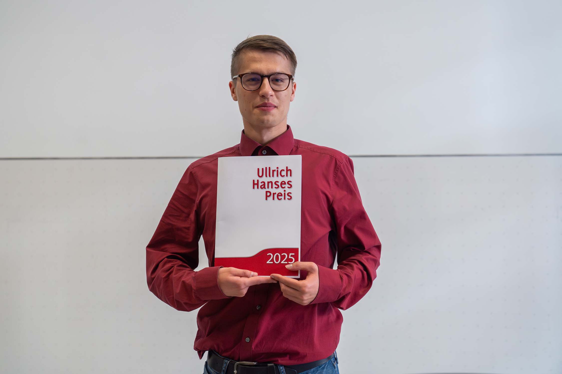 Vor einem schlichten Hintergrund steht eine Person mit Brille und rotem Hemd und hält eine weiß-rote Urkunde mit der Aufschrift „Ullrich Hanses Preis 2025“, mit der mit den Hanses-Preisen Leistungen im Bauingenieurwesen gewürdigt werden. at Hochschule Coburg