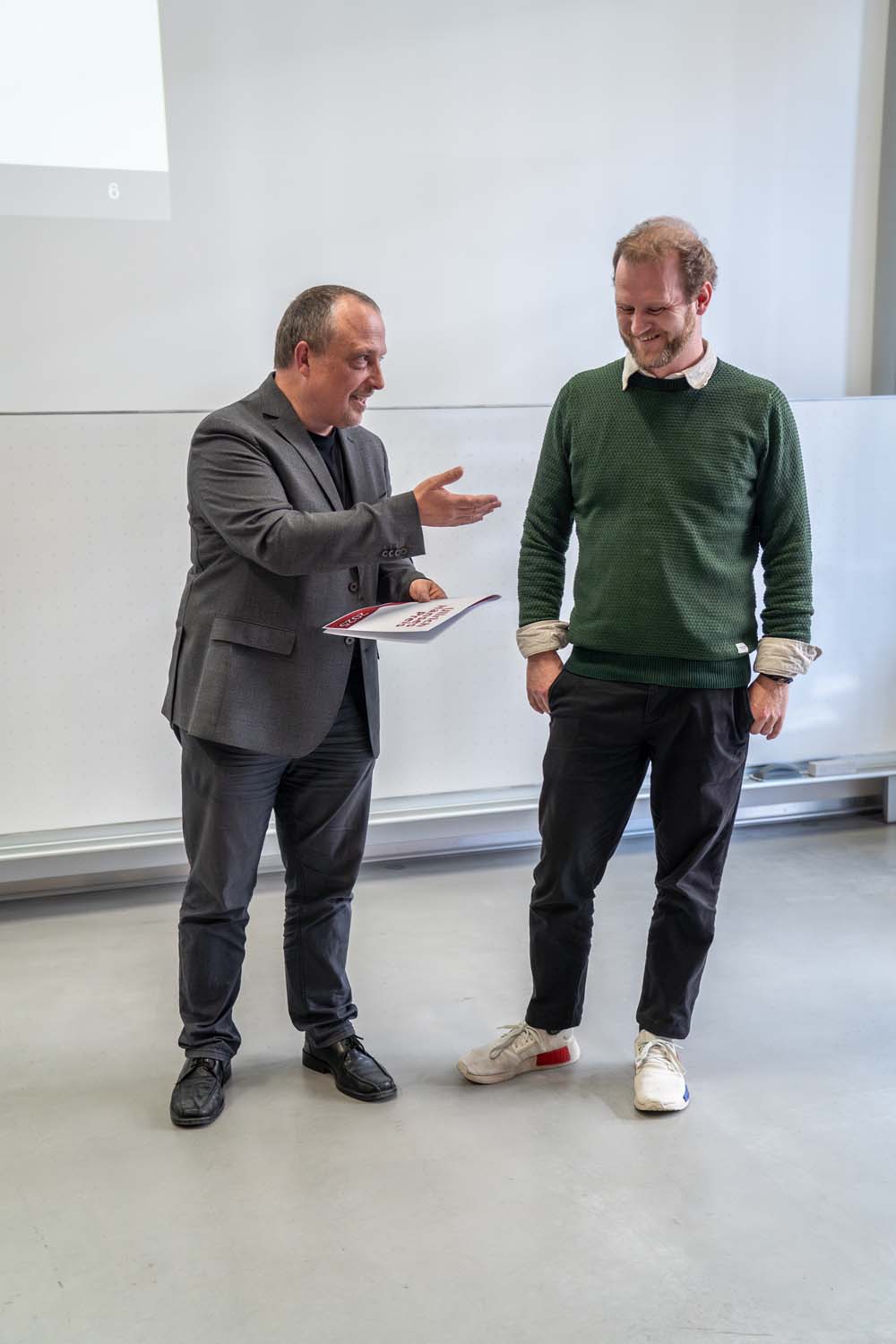 Zwei Männer stehen in einem Gebäude auf grauem Boden. Der linke Mann, der einen grauen Anzug trägt, gestikuliert mit einer Hand und hält einen Ordner – möglicherweise für die Kurbasik- und Hanses-Preise im Bauingenieurwesen. Der rechte Mann, der einen grünen Pullover und weiße Turnschuhe trägt, lächelt und blickt nach unten. at Hochschule Coburg