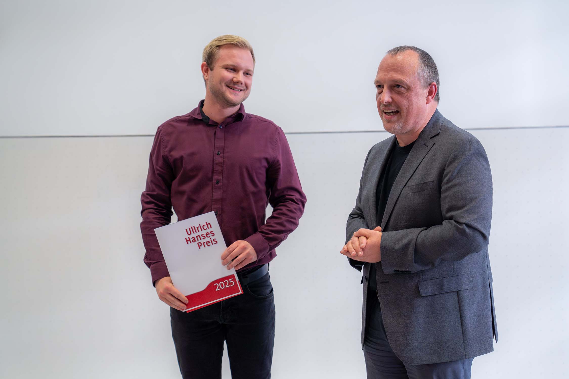 In einem Gebäude stehen zwei Männer nebeneinander, der eine hält lächelnd eine Broschüre mit der Aufschrift „Kurbasik- und Hanses-Preise 2025“, der andere gestikuliert angeregt – beide unterhalten sich über Nachwuchs im Bauingenieurwesen. at Hochschule Coburg