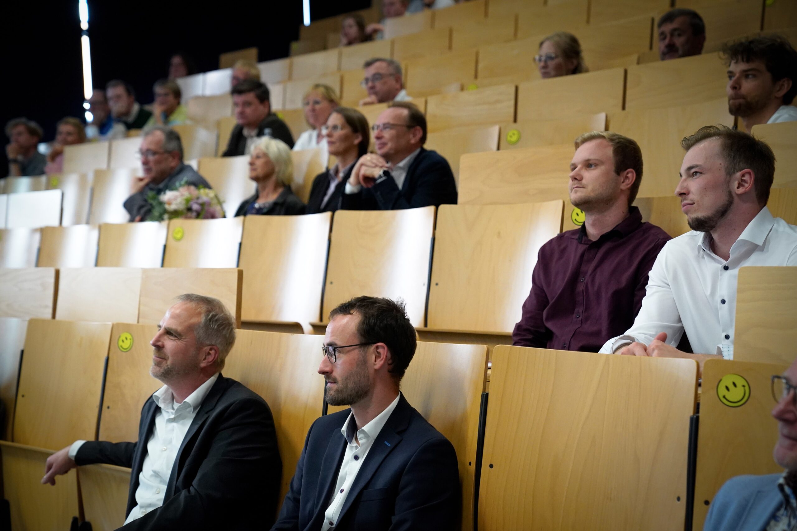 Menschen sitzen in einem Hörsaal mit Holzsitzen, manche mit Smiley-Aufklebern. Die meisten beobachten oder hören aufmerksam etwas außerhalb ihres Sichtfelds. Die allgemeine Stimmung ist formell und konzentriert. at Hochschule Coburg
