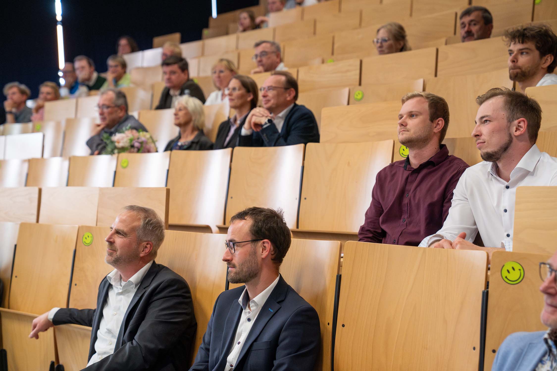 Menschen sitzen in einem Hörsaal mit Holzsitzen, einige davon mit gelben Smiley-Aufklebern dekoriert. Die meisten Teilnehmer, vermutlich aus dem Bauingenieurwesen oder aus dem Bereich Kurbasik-Preise, wirken aufmerksam und interessiert und blicken nach vorne zum Redner. at Hochschule Coburg