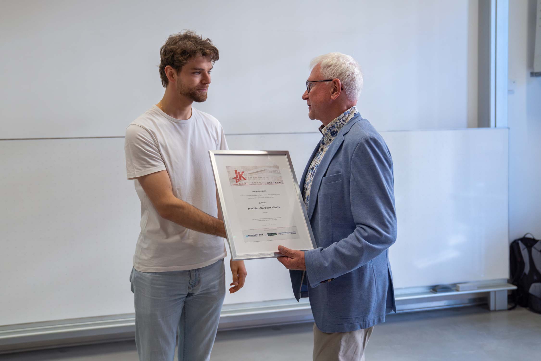 Ein junger Mann im weißen T-Shirt schüttelt einem älteren Mann im hellblauen Blazer die Hand. Er hält eine gerahmte Kurbasik-Preis-Urkunde in der Hand. Sie stehen drinnen vor einer weißen Tafel. at Hochschule Coburg