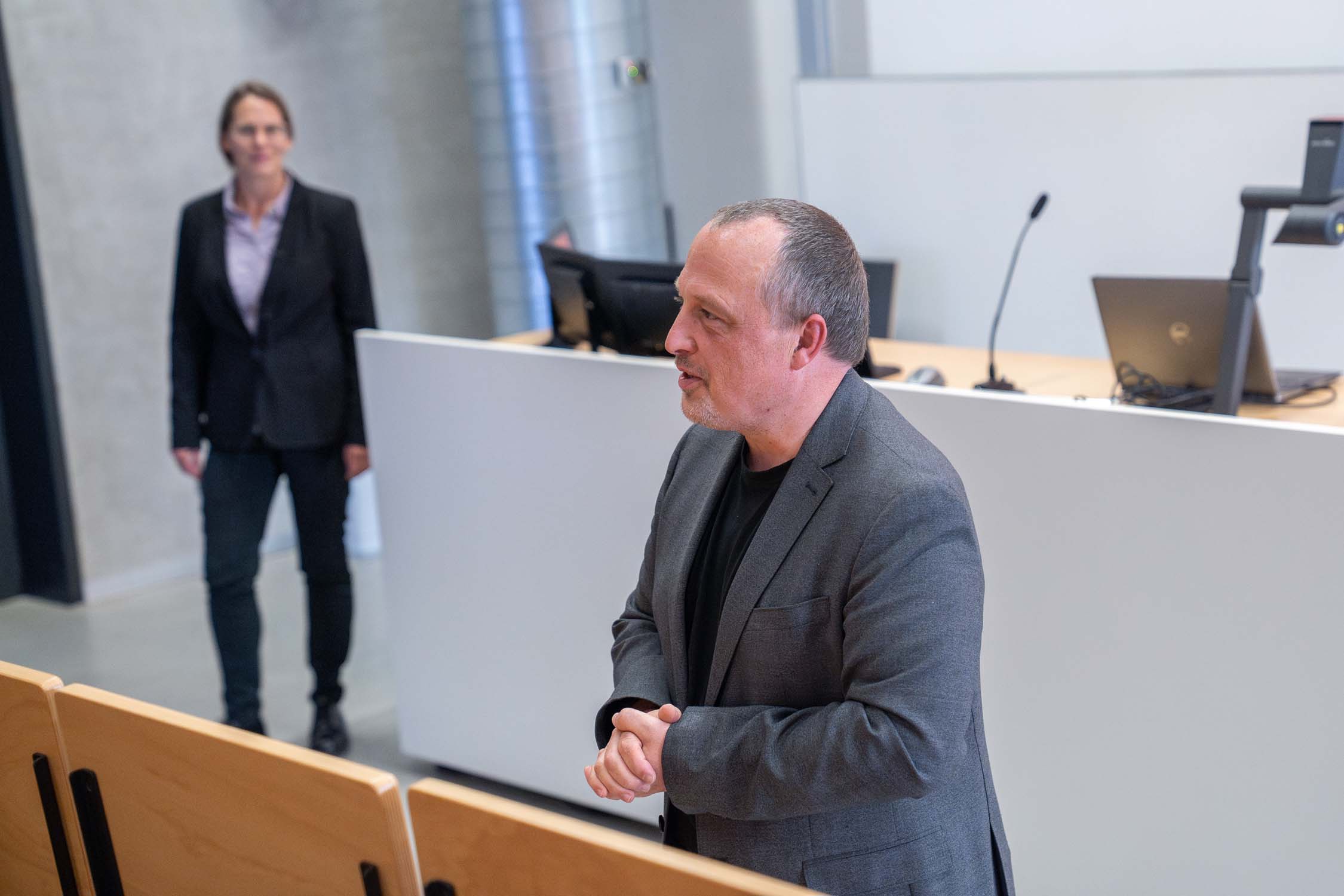 Ein Mann im grauen Blazer spricht bei einer Veranstaltung des Fachbereichs Bauingenieurwesen in einem modernen Hörsaal vor Publikum. In der Nähe des Podiums steht eine Frau – vielleicht wartet sie auf die Bekanntgabe der Gewinner des Nachwuchs- oder Kurbasik- und Hanses-Preises. at Hochschule Coburg