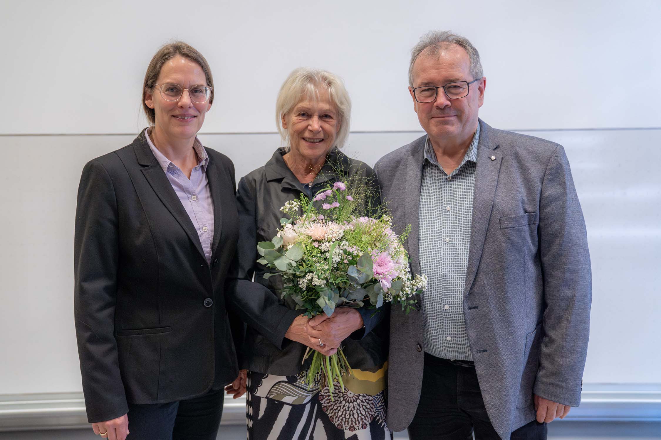 Drei Erwachsene stehen lächelnd nebeneinander. Die Frau in der Mitte, die einen Blumenstrauß hält, scheint eine Kurbasik-Preisträgerin zu sein. Alle sind in Business- oder halbformeller Kleidung gekleidet und posieren vor einer weißen Wand. at Hochschule Coburg