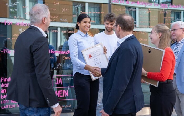 Eine Frau lächelt, als sie während einer Veranstaltung im Freien von einem älteren Herrn eine Urkunde für den Kurbasik- und Hanses-Preis 2025 entgegennimmt. Mehrere Personen stehen um sie herum, einige halten Urkunden in den Händen, auf der Glaswand dahinter steht in rosa „Nachwuchs“. at Hochschule Coburg