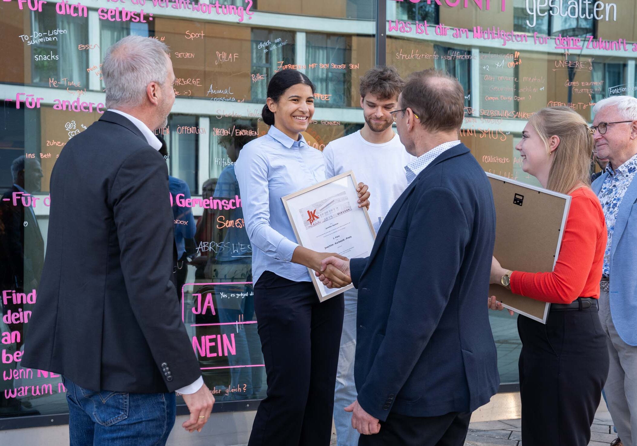 Eine Frau lächelt, als sie während einer Veranstaltung im Freien von einem älteren Herrn eine Urkunde für den Kurbasik- und Hanses-Preis 2025 entgegennimmt. Mehrere Personen stehen um sie herum, einige halten Urkunden in den Händen, auf der Glaswand dahinter steht in rosa „Nachwuchs“. at Hochschule Coburg