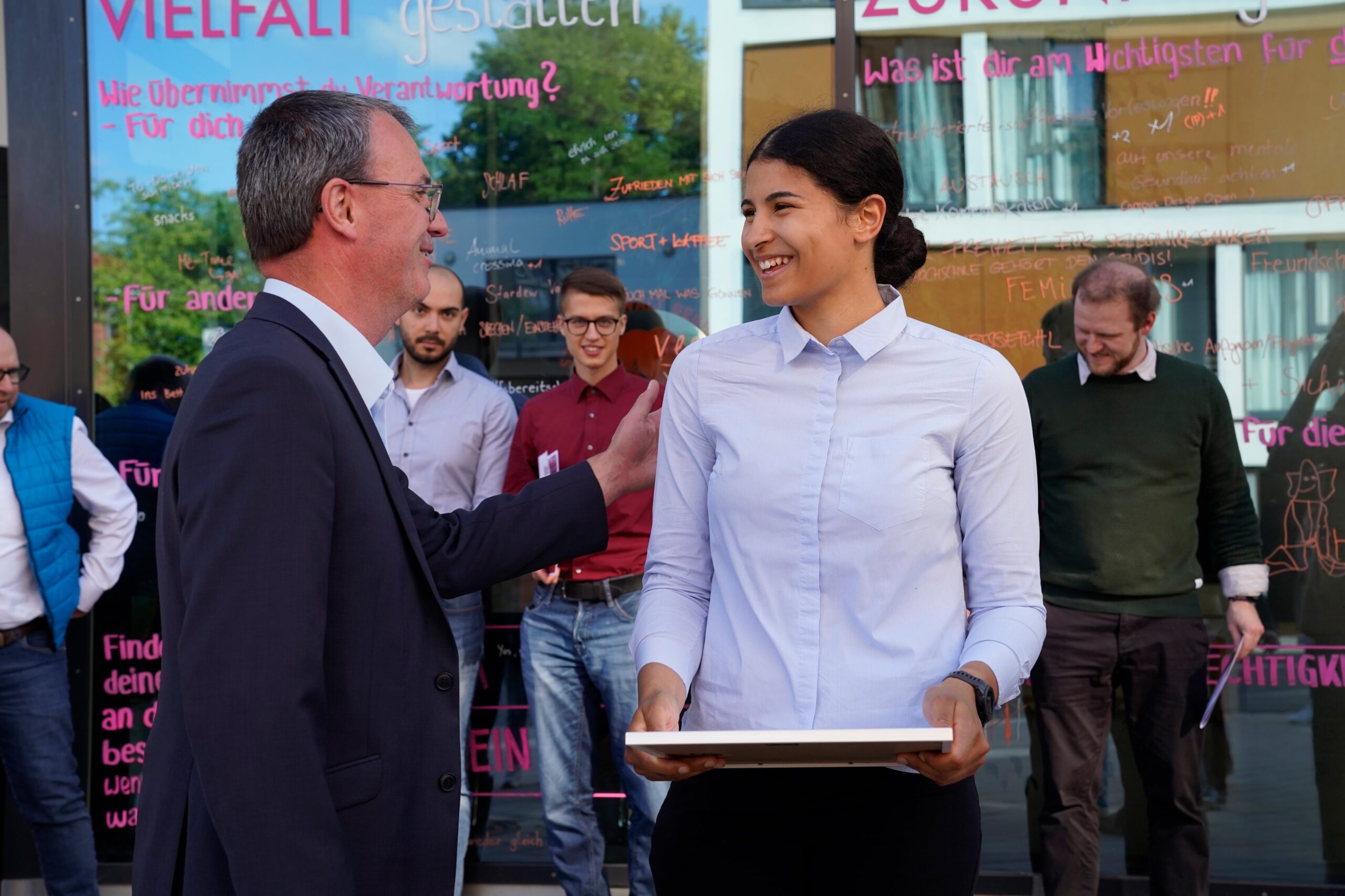 Ein Mann im Anzug spricht mit einer lächelnden Frau, die einen Laptop hält, während im Hintergrund mehrere Personen vor einem Gebäude mit bunten Schriftzügen an den Fenstern stehen. at Hochschule Coburg