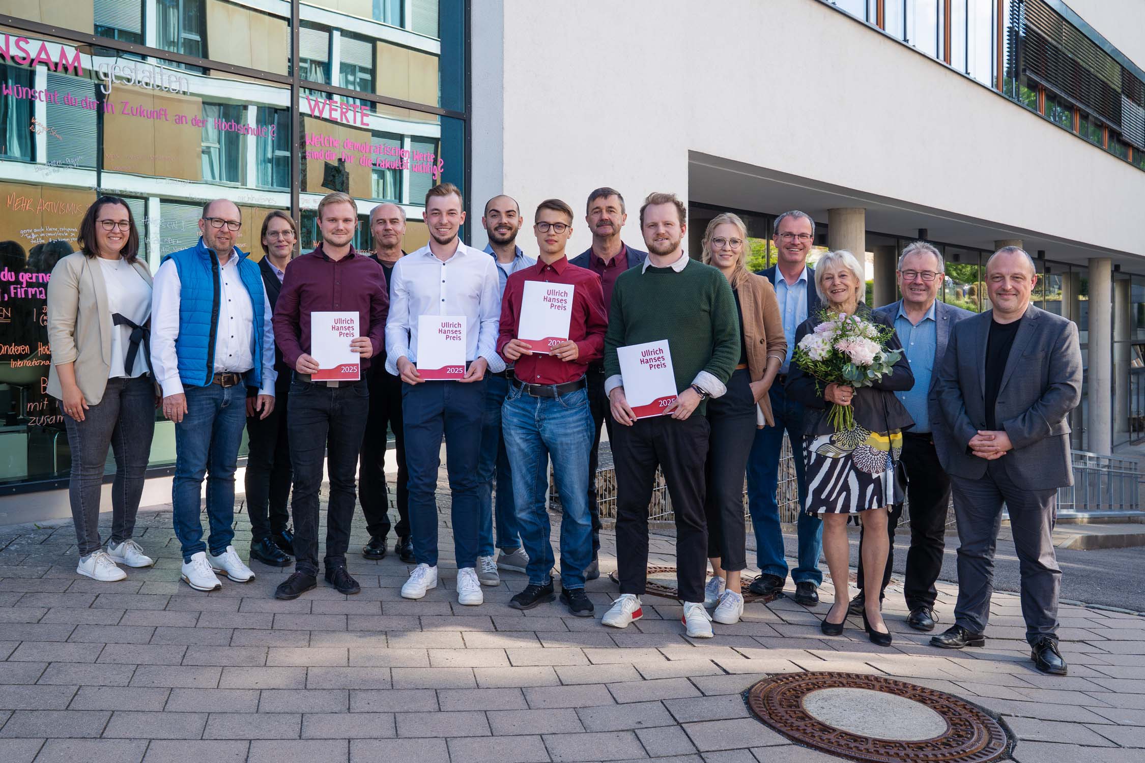Eine Gruppe von fünfzehn Personen steht lächelnd vor einem Gebäude und feiert die Verleihung des Kurbasik- und Hanses-Preises. Vier Personen vorn halten Urkunden mit Auszeichnungen für ihre Leistungen im Bauingenieurwesen. Eine Frau hält einen Blumenstrauß; dahinter spiegeln sich Glasfenster mit Text. at Hochschule Coburg