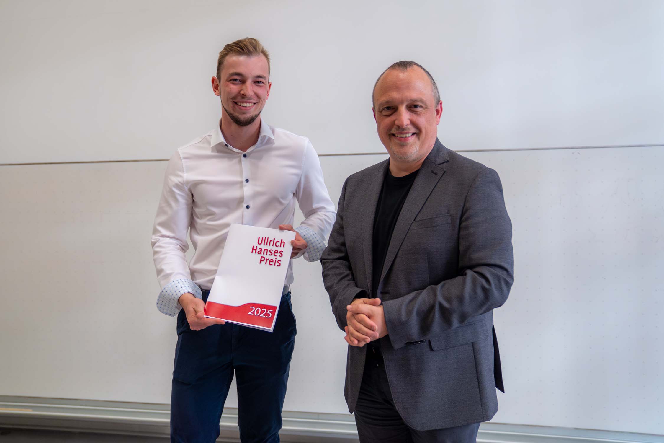 Zwei Männer stehen lächelnd im Innenbereich; einer hält einen weiß-roten Ordner mit der Aufschrift „Ulrich-Hanses-Preis 2025“. Beide, in Business-Kleidung, feiern vor einer weißen Wand ihre Erfolge im Bauingenieurwesen. at Hochschule Coburg