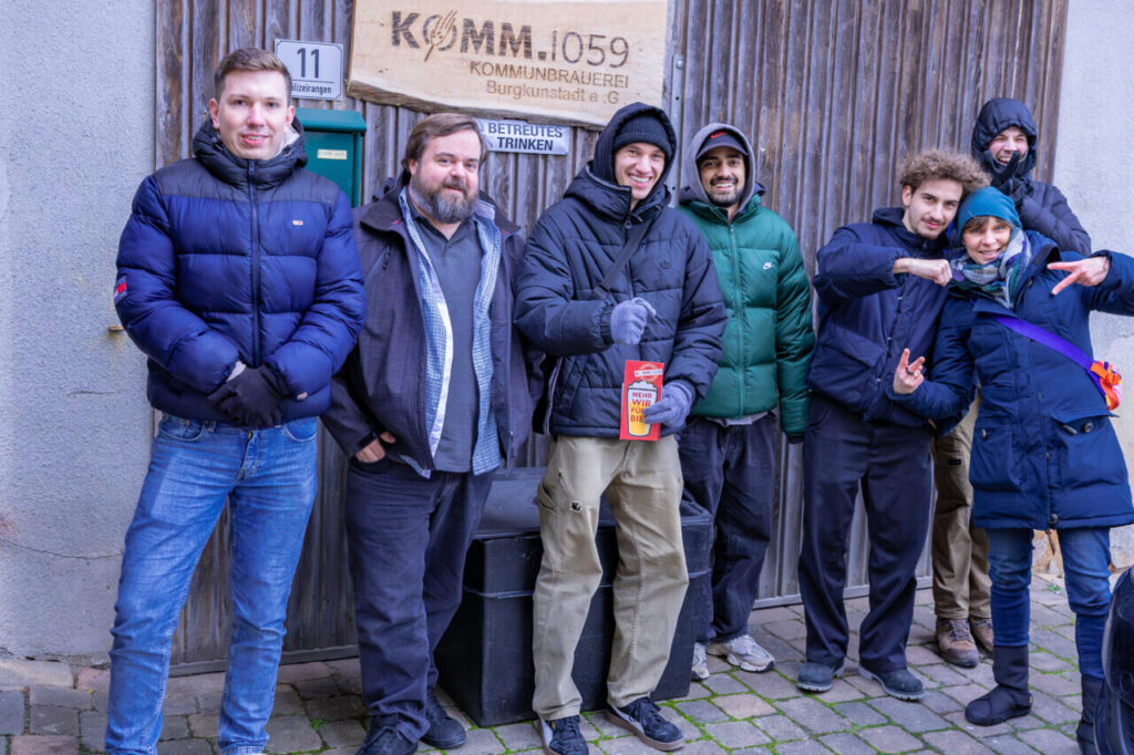 Eine Gruppe von sieben Personen in Winterkleidung, möglicherweise Studierende der Wirtschaftsinformatik 2.0, posiert lächelnd vor einer Holztür mit einem „KOMM.1059“-Schild. Der Kopfsteinpflasterboden verleiht ihr Charme, während einige Peace-Zeichen zeigen oder verspielt gestikulieren. at Hochschule Coburg