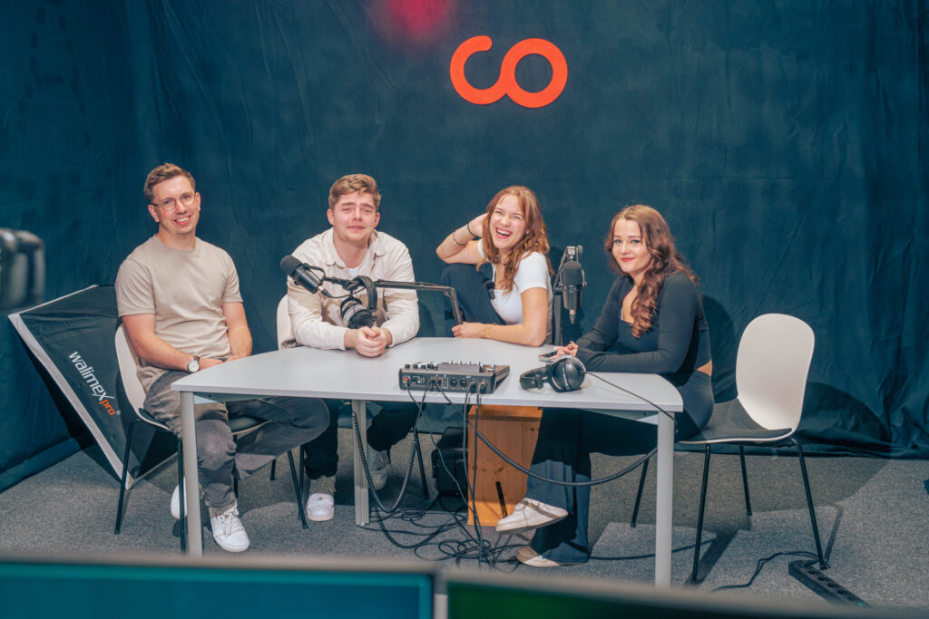 In einem Podcast-Studio mit dunklem Hintergrund und orangefarbenem Unendlichkeitssymbol sitzen vier junge Erwachsene an einem Tisch mit Mikrofonen und Audio-Equipment und lächeln in die Kamera – bereit, über Wirtschaftsinformatik 2.0 zu diskutieren. at Hochschule Coburg