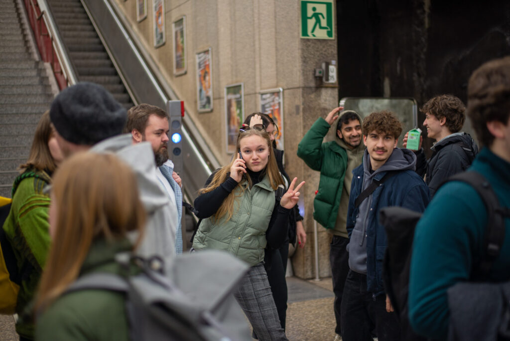 Eine junge Frau steht lächelnd in der Mitte einer Gruppe, hält ein Telefon ans Ohr und macht ein Peace-Zeichen – vielleicht feiert sie damit ihren neuesten Erfolg in Wirtschaftsinformatik 2.0. Die Leute um sie herum unterhalten sich in der Nähe einer Rolltreppe und eines Notausgangsschilds. at Hochschule Coburg