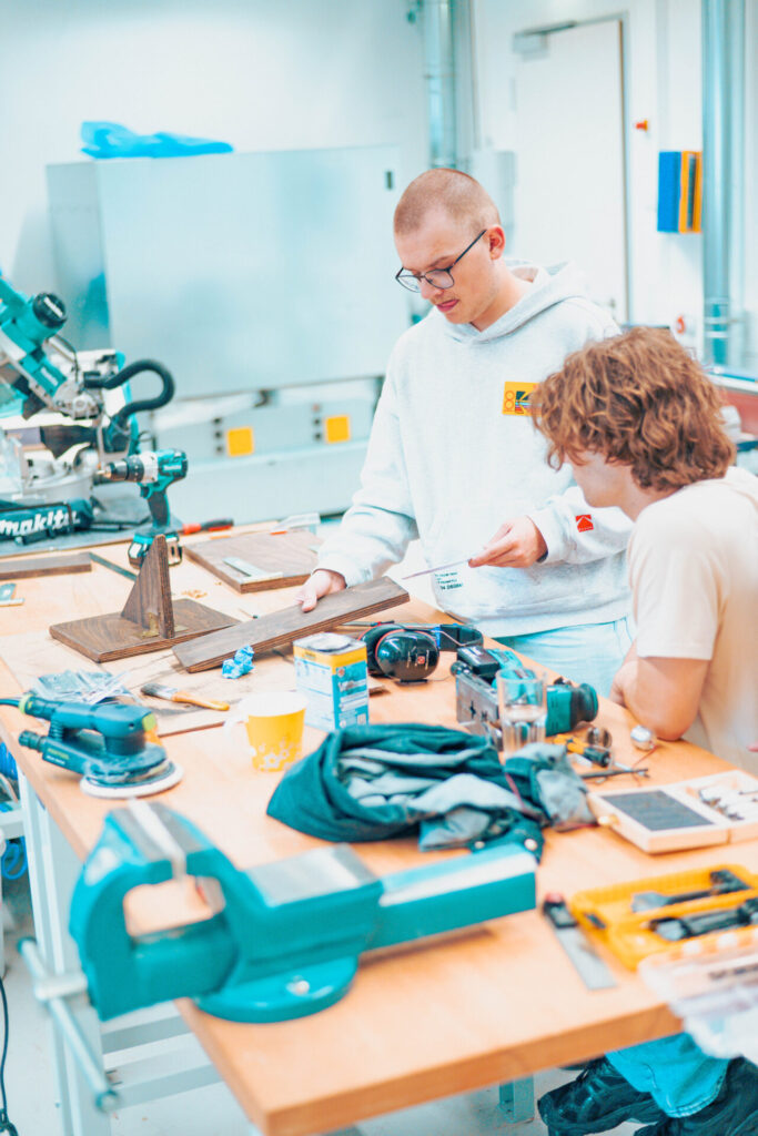 Zwei junge Leute arbeiten gemeinsam an einem überfüllten Werkstatttisch und wenden Konzepte der Wirtschaftsinformatik an, um Werkzeuge und Holzstücke zu organisieren, während sie sich in einer hellen, modernen Werkstatt auf ein Holzbearbeitungsprojekt konzentrieren. at Hochschule Coburg