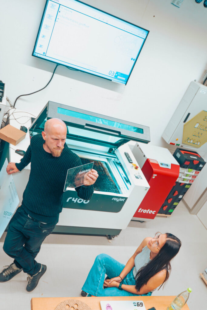 Ein Mann steht mit einem durchsichtigen Blatt in der Hand da und erklärt einer Frau etwas über Wirtschaftsinformatik. Sie sitzt neben einer Laserschneidmaschine in einem modernen Arbeitsbereich, hinter dem ein großer Bildschirm technische Diagramme anzeigt. at Hochschule Coburg
