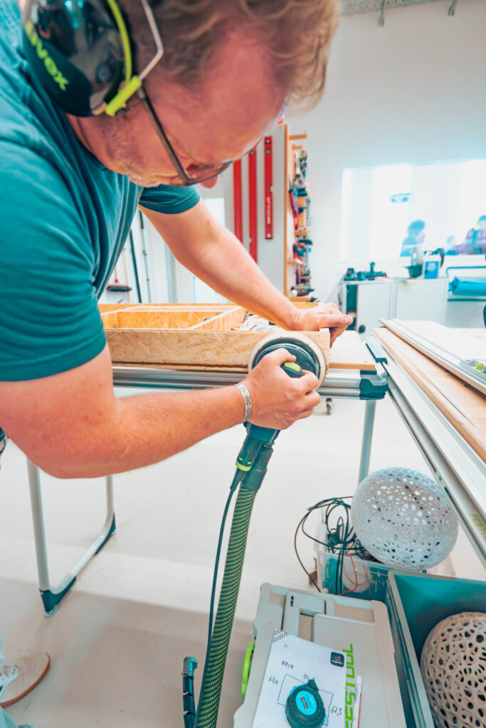 Eine Person mit Gehörschutz bedient einen elektrischen Schleifer auf einer Holzplatte in einer hellen Werkstatt, in der Werkzeuge und Materialien geordnet sind – ein Spiegelbild der Effizienz der Wirtschaftsinformatik 2.0 bei der Verwaltung von Prozessen und der Organisation von Arbeitsbereichen. at Hochschule Coburg